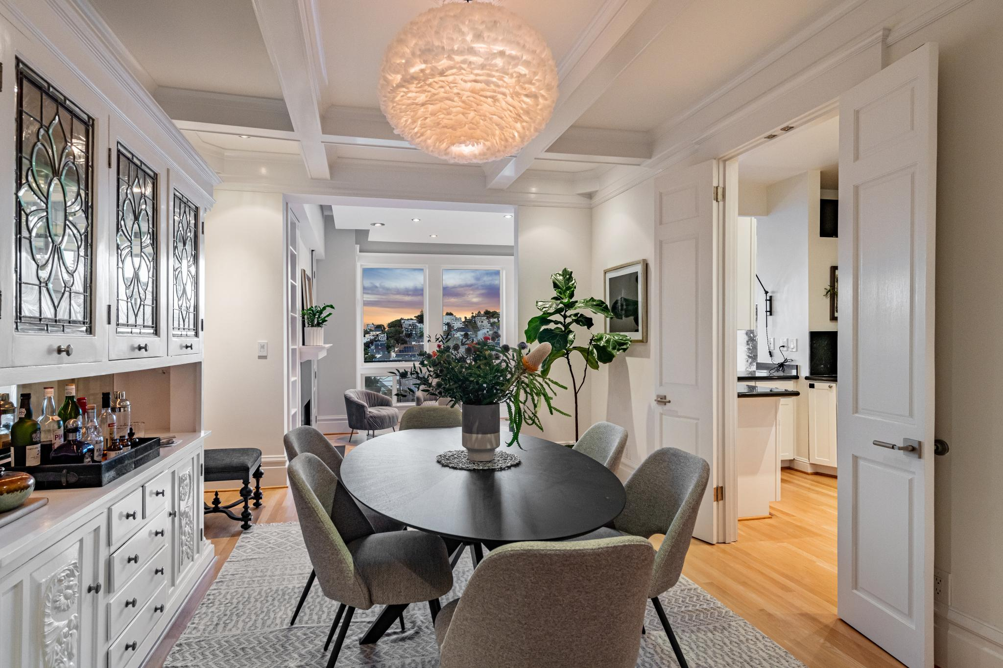 A cozy dining room features a round table with six chairs, a modern chandelier, a built-in cabinet with stained glass, and a view into a bright living area and kitchen.