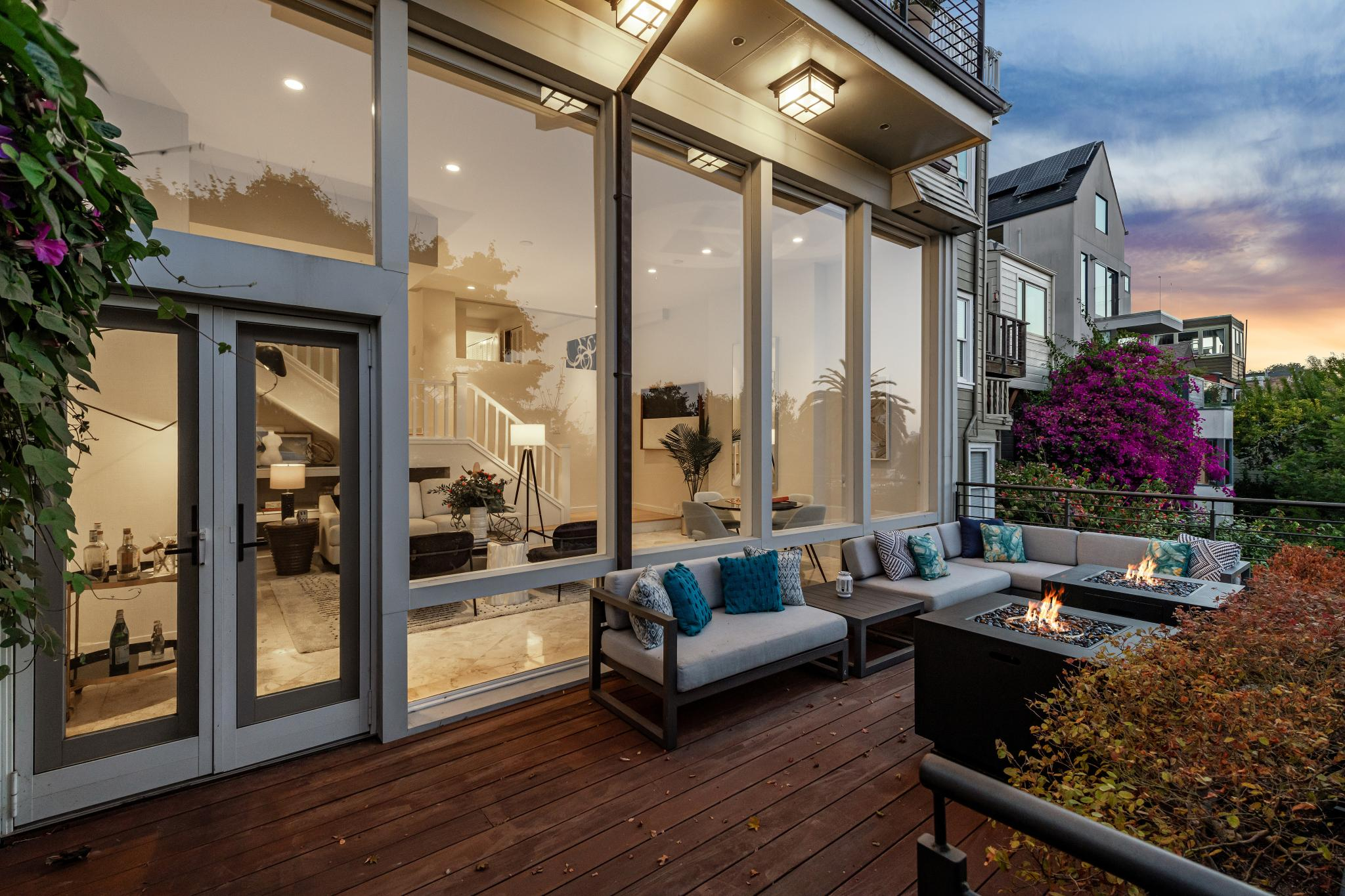 A cozy outdoor patio with a large sectional sofa, fire pit tables, and lush greenery, connected to a modern interior through glass doors and large windows at dusk.