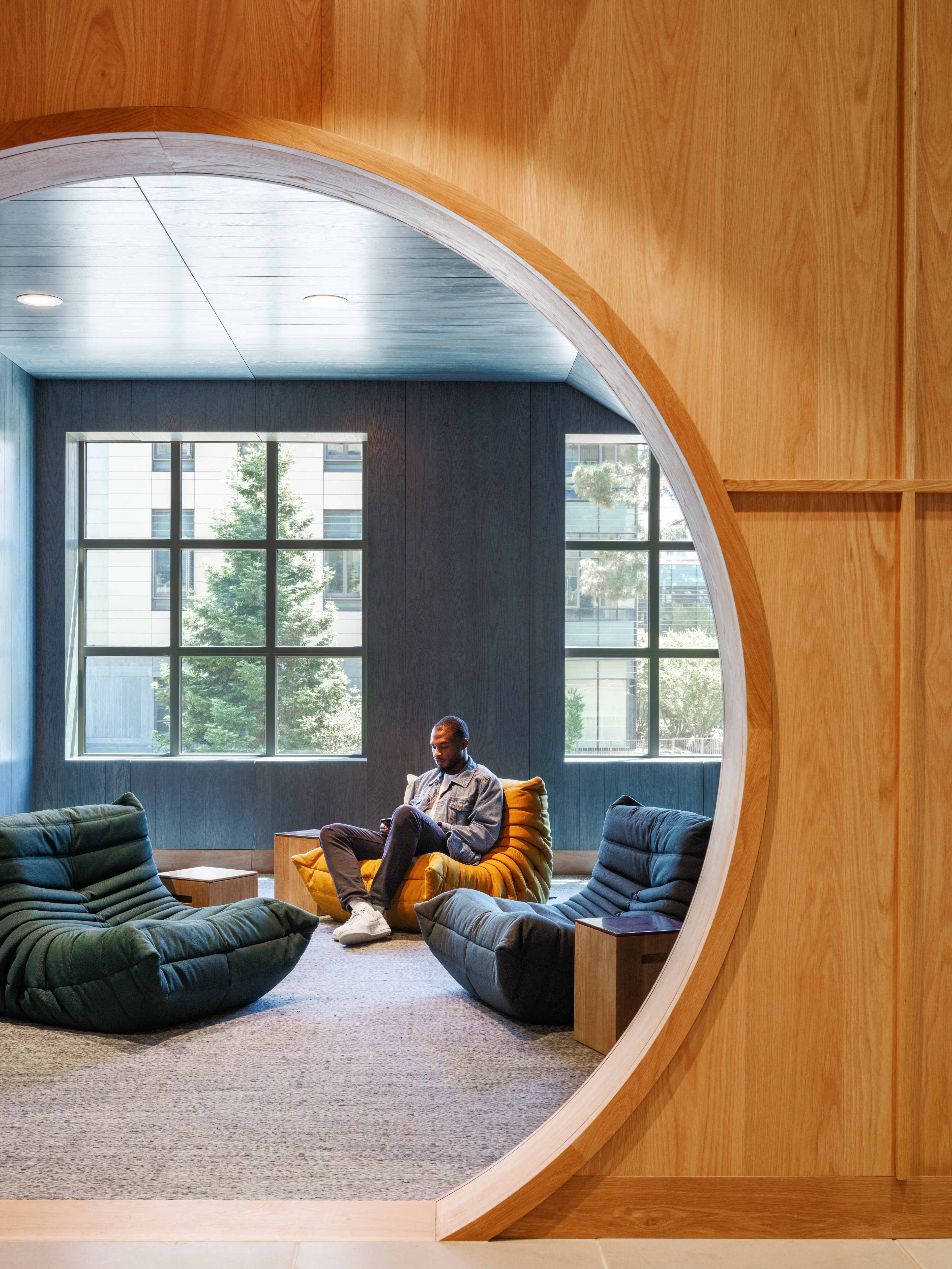 A man sits on a yellow plush chair in a cozy, modern lounge with large windows, flanked by green and gray chairs. The arched wooden doorway frames the scene.