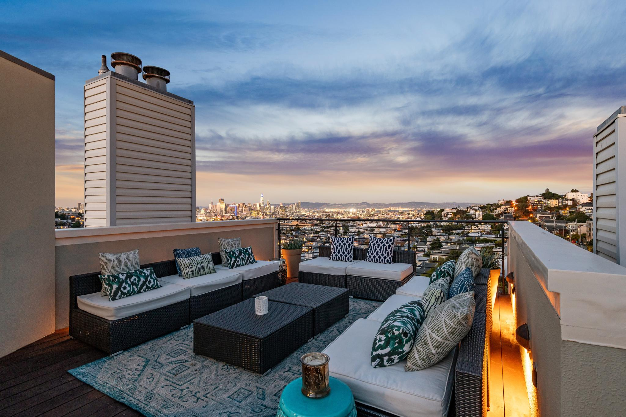 A rooftop patio with cushioned seating, decorative pillows, and a central table overlooks a cityscape at sunset, with a mix of buildings and hills in the background.