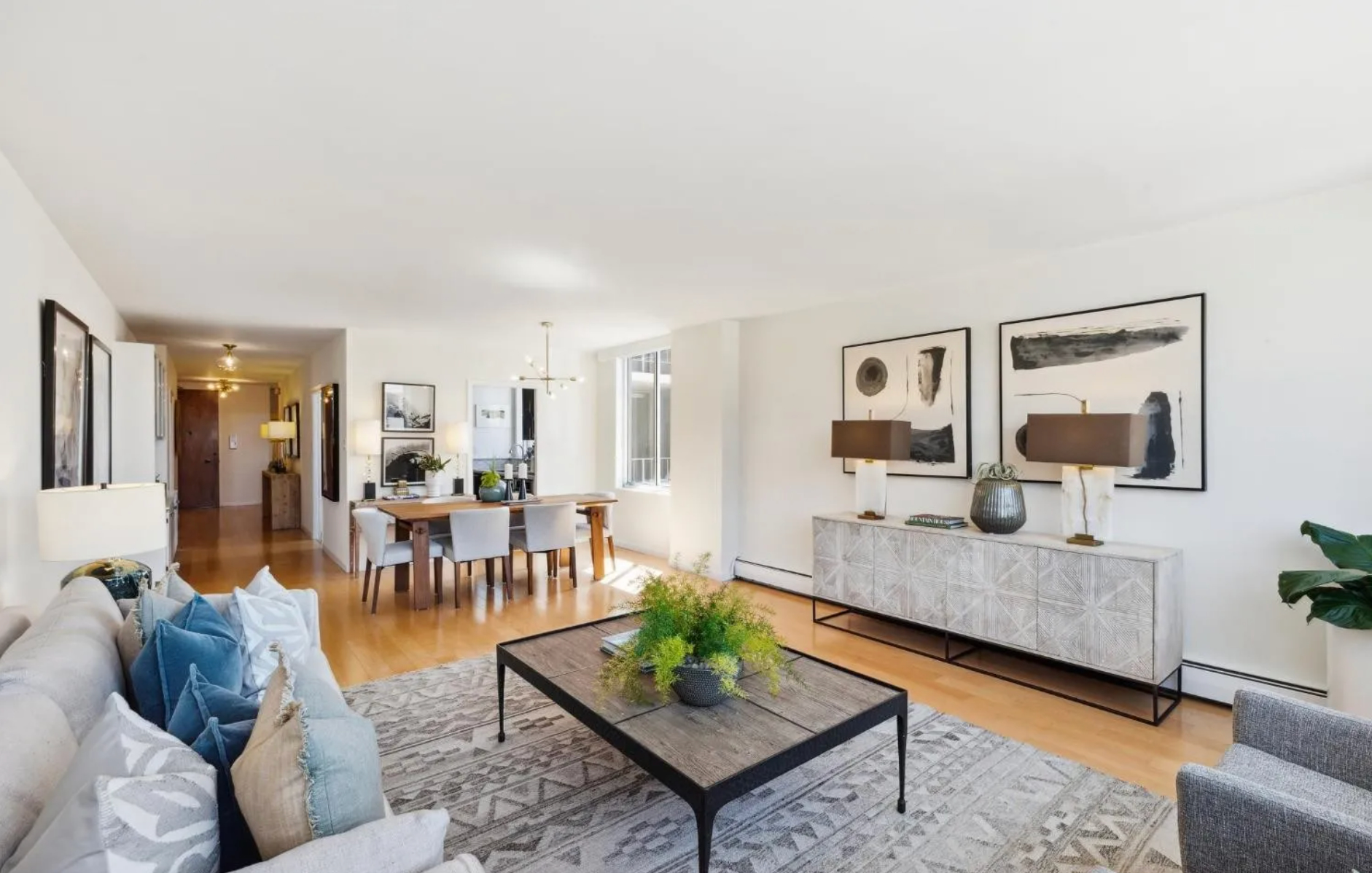 A contemporary living and dining area featuring light wood floors, a beige sofa with blue and gray pillows, a modern coffee table with plants, and abstract wall art.