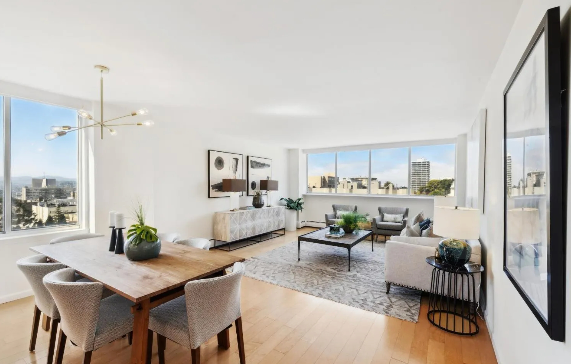 A modern, sunlit living-dining area with large windows showing a city view. It has a wooden dining table, plush chairs, and a cozy seating area with a rug and coffee table.