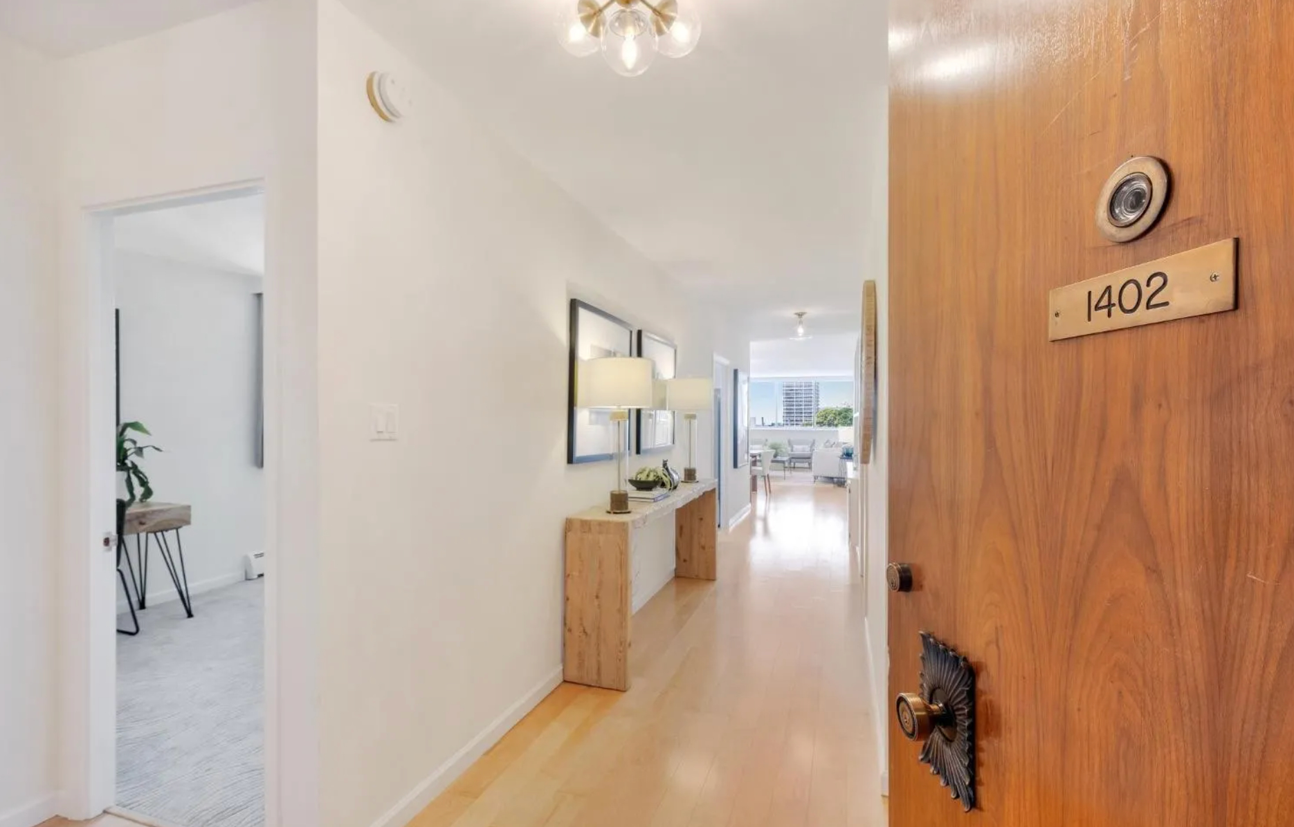 The image shows an open wooden door marked &quot;1402&quot; leading to a well-lit hallway with wooden flooring. A side room is visible on the left, and the hallway leads to a cozy living space.