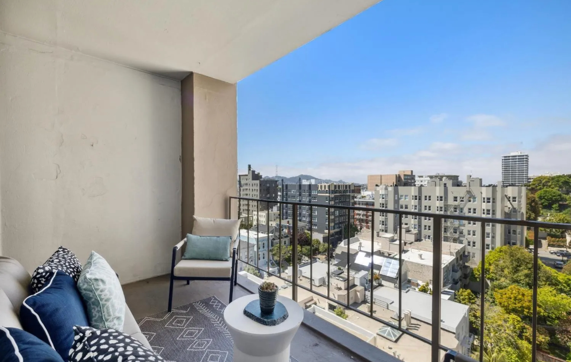 A cozy balcony with outdoor seating, including a sofa and chair with blue and white cushions, a small table, and a view of city buildings and clear blue sky.