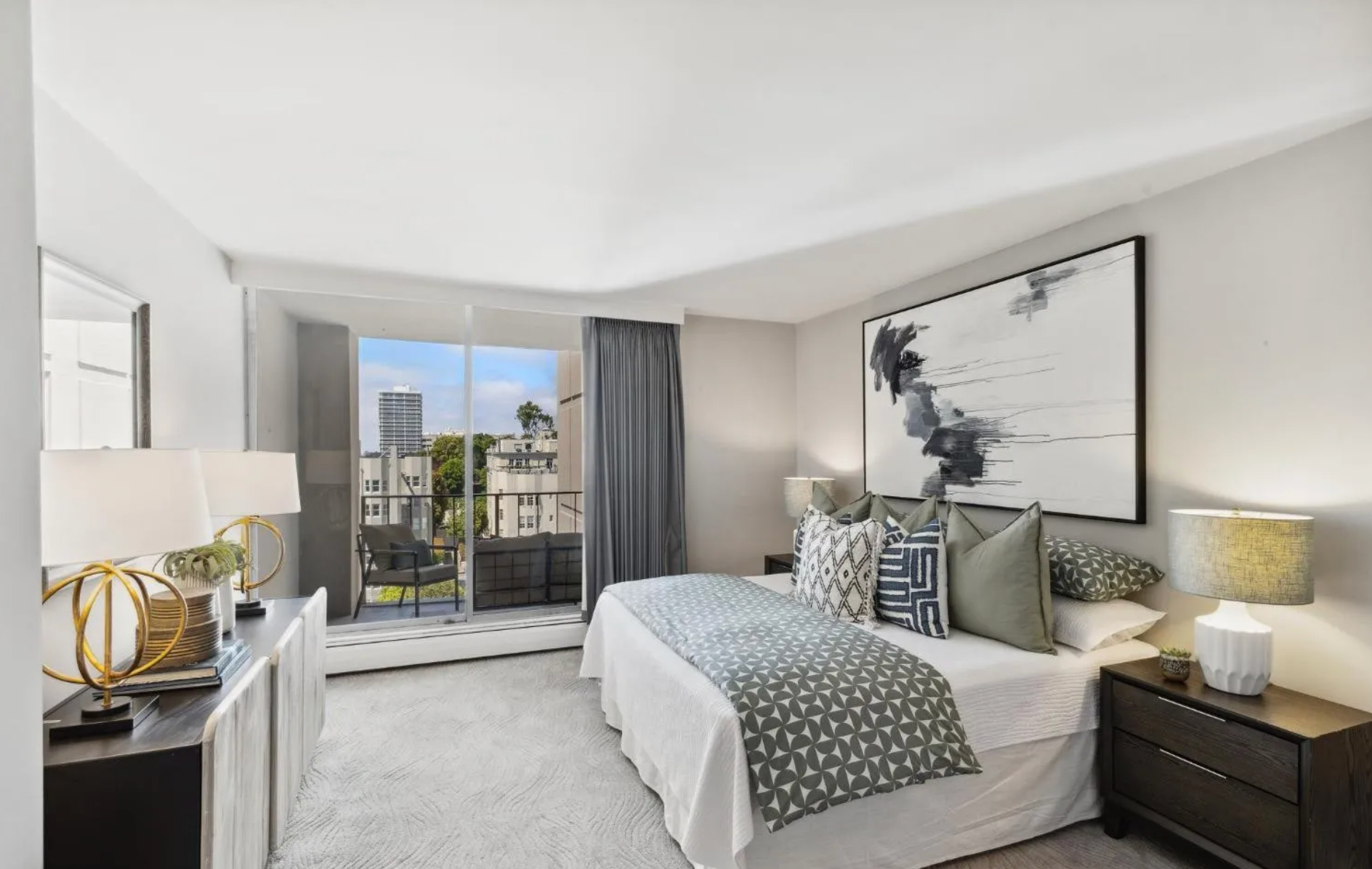 The image shows a modern bedroom with large windows, a double bed with geometric-patterned bedding, two bedside tables with lamps, and abstract artwork above the bed.