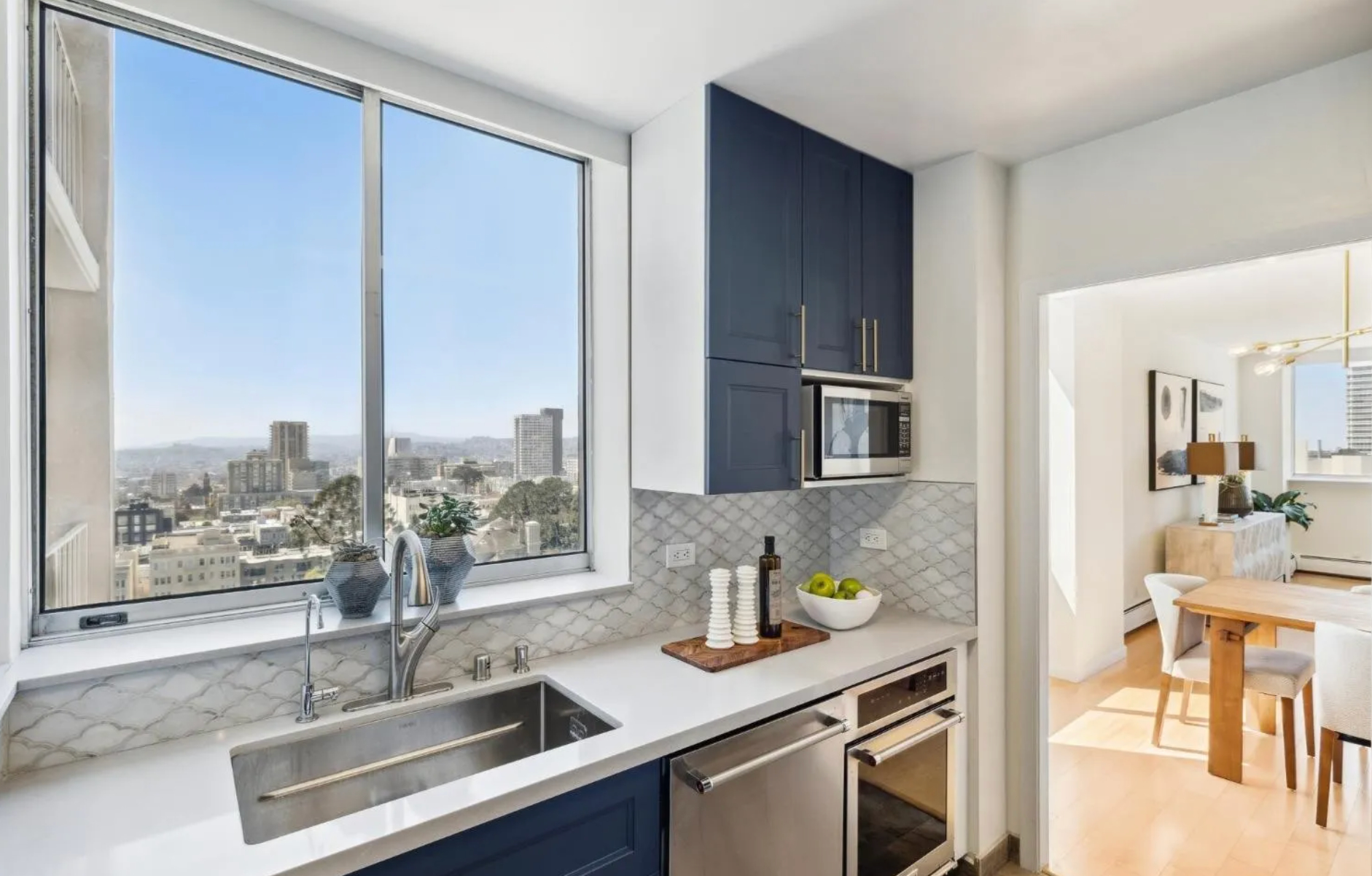 A modern kitchen with blue cabinets, a sink, and a city view window leads to a bright dining and living area with light wood flooring and stylish decor.