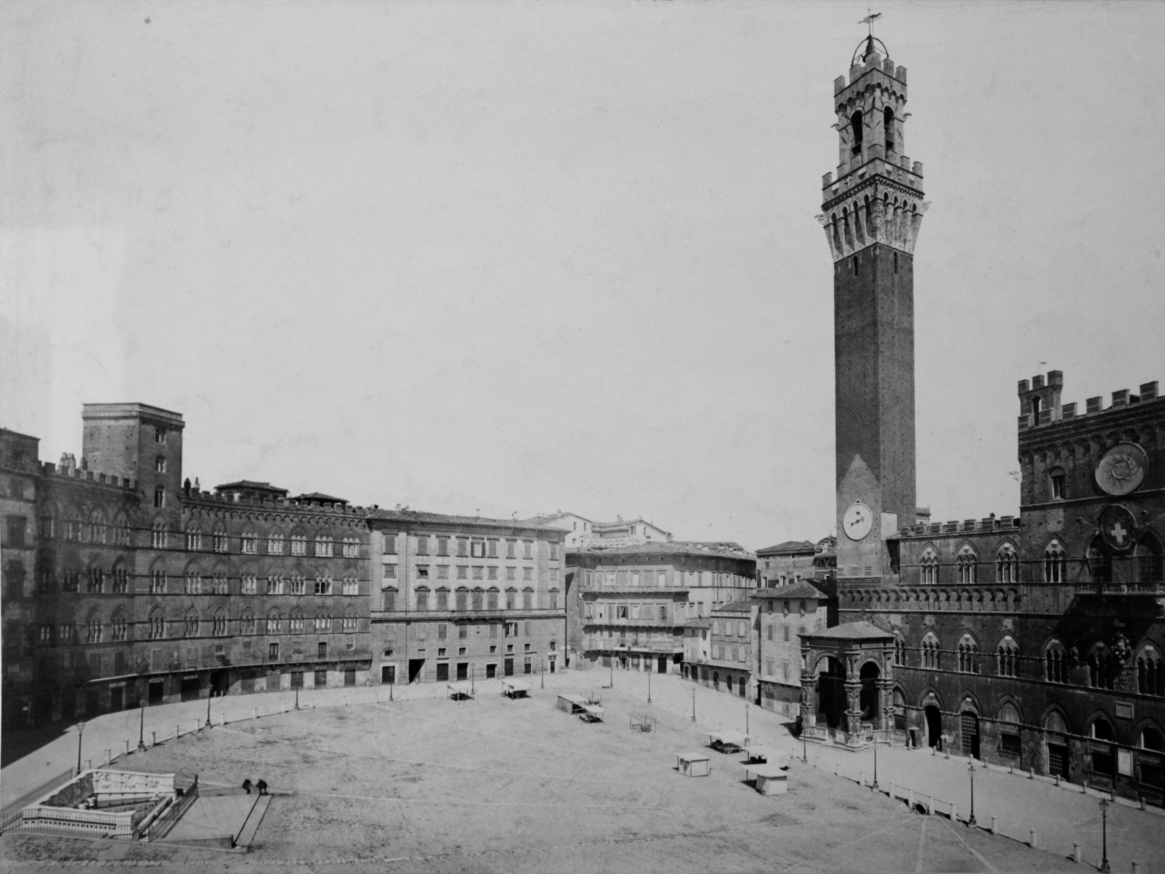 The image shows a large, empty medieval square with surrounding multi-story buildings and a tall clock tower with battlements on one side, typical of historic Italian architecture.