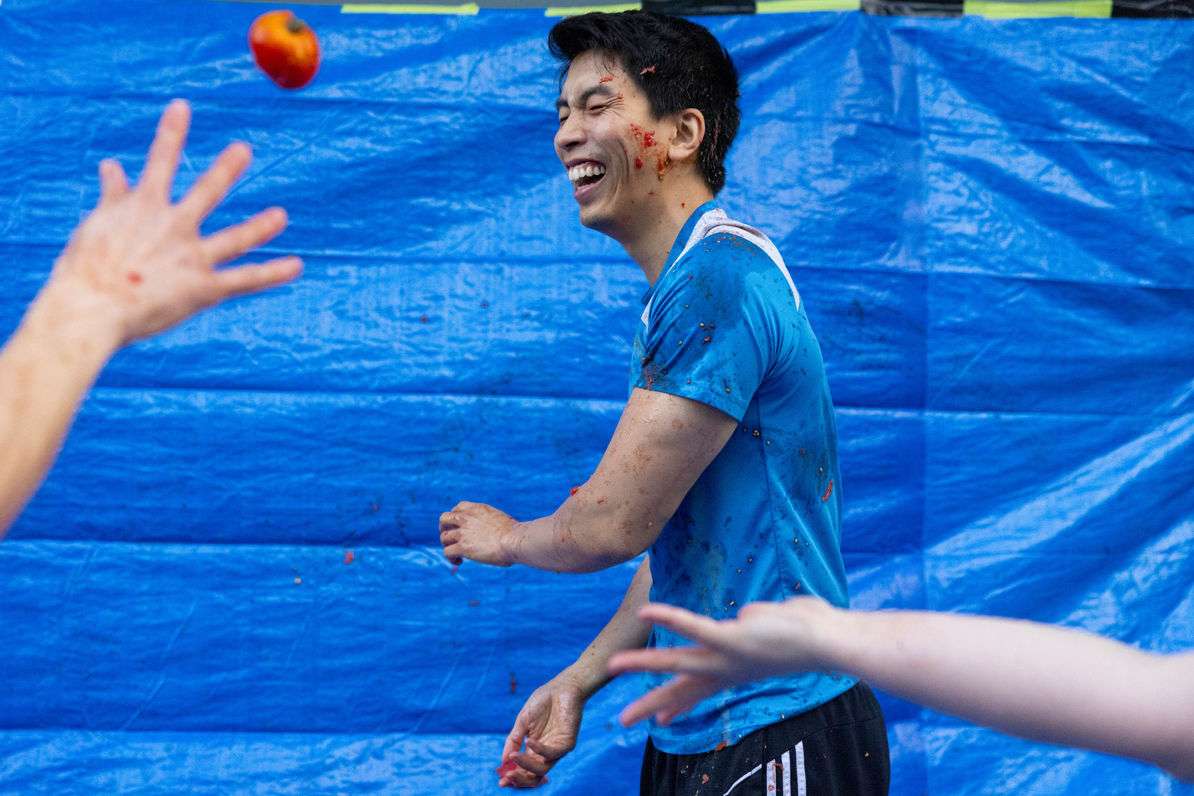 A person in a blue shirt smiles while engaging in a playful activity involving throwing tomatoes, with a blue tarp as a backdrop.