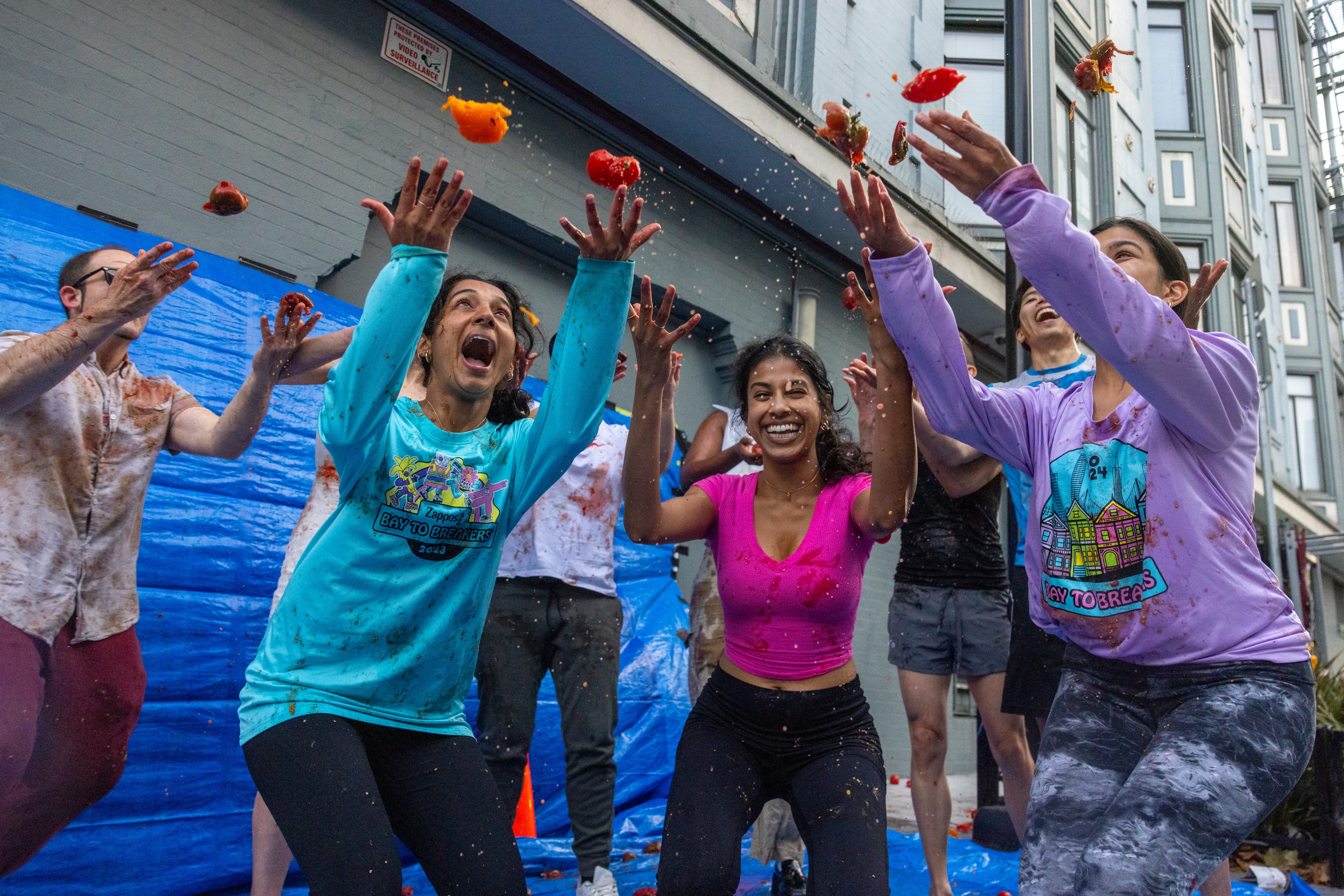 People are joyfully throwing tomatoes in an outdoor setting, splattering juice. They appear excited and are wearing casual clothes, some with logos, in a blue-tarp area.