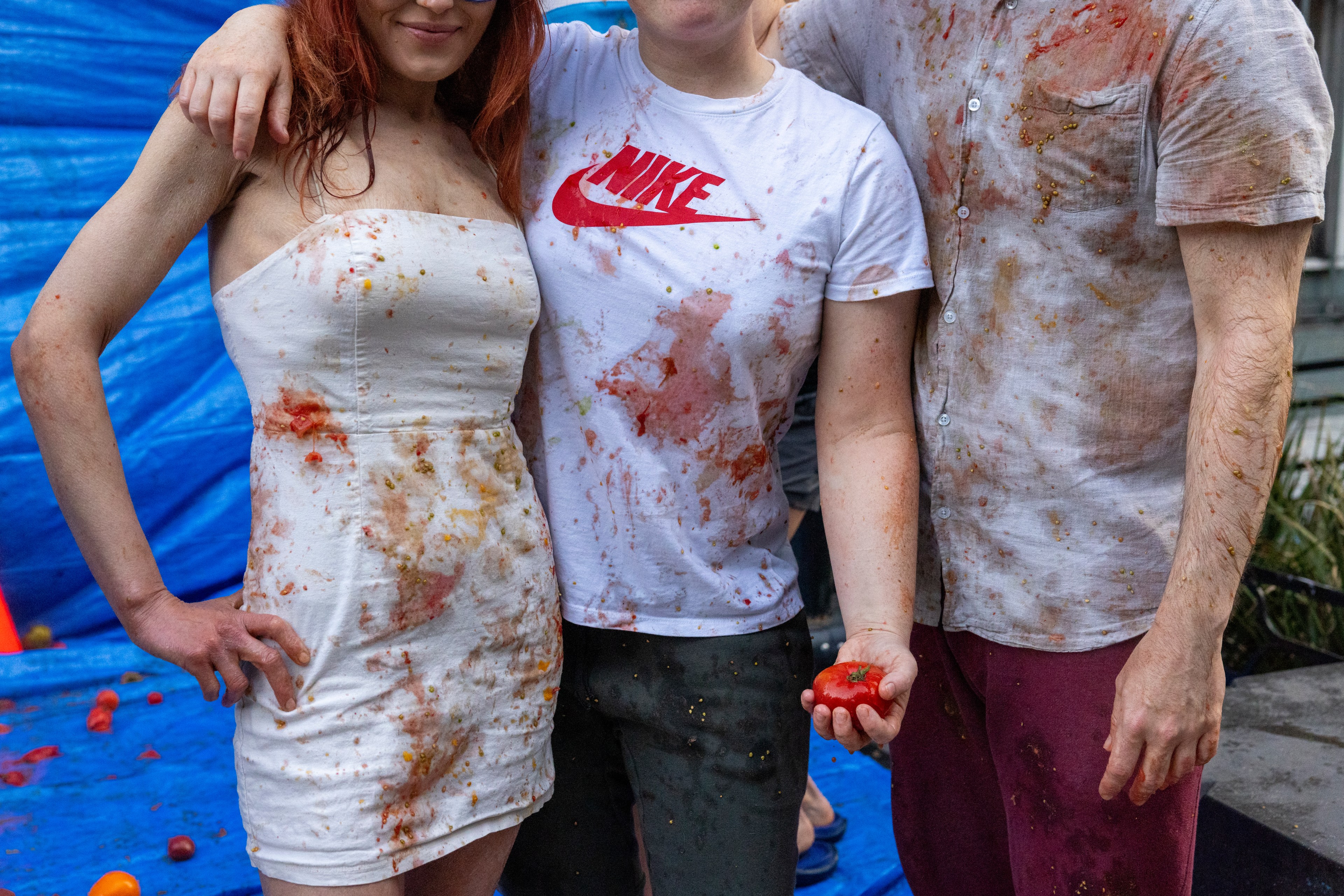 Three people, each wearing thoroughly soiled clothing, pose in front of a blue tarp, likely after a messy event involving tomatoes, as one holds a tomato in hand.