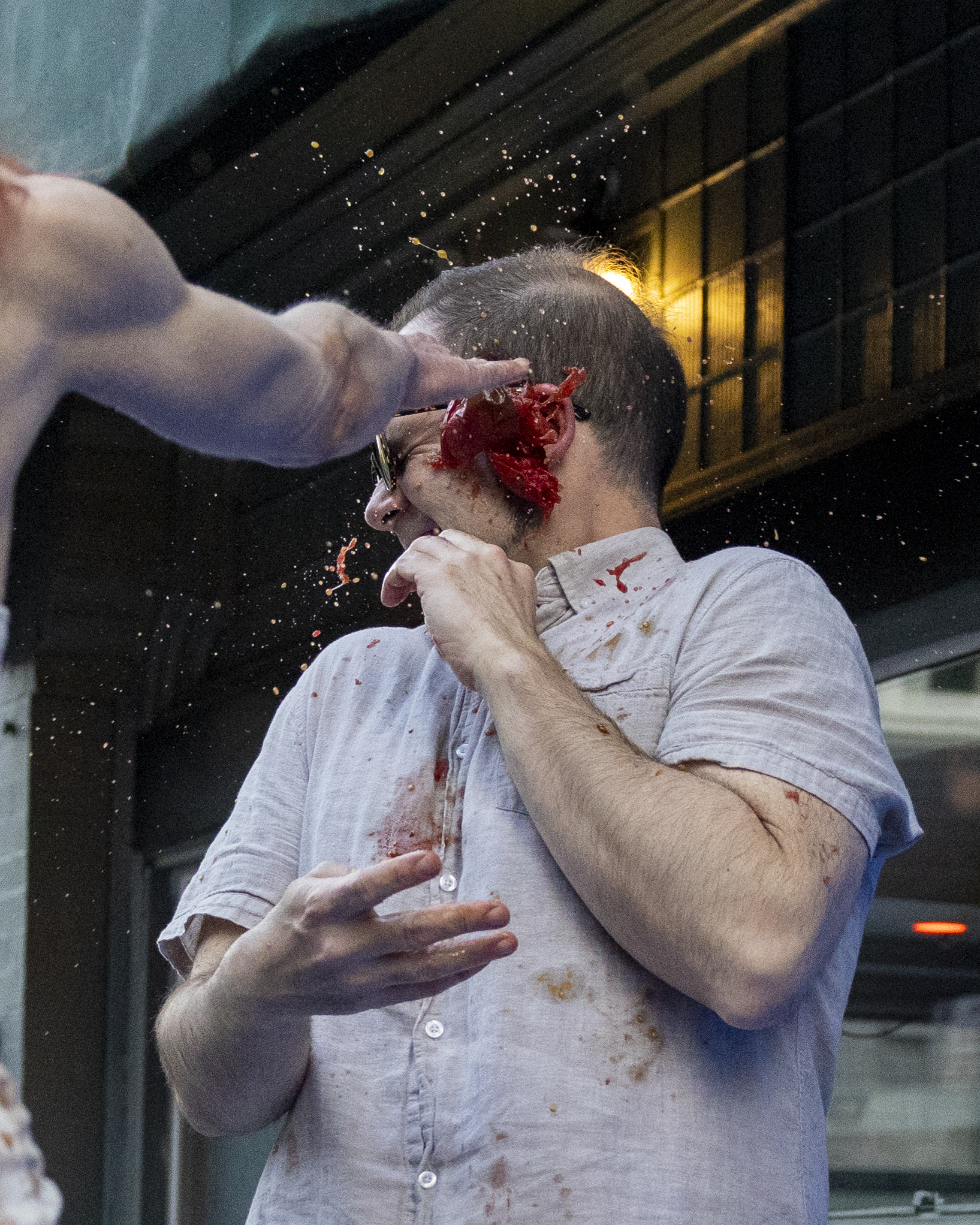 A man in a light-colored shirt and glasses is being hit in the face by a flying tomato, causing juice and pulp to splash everywhere.