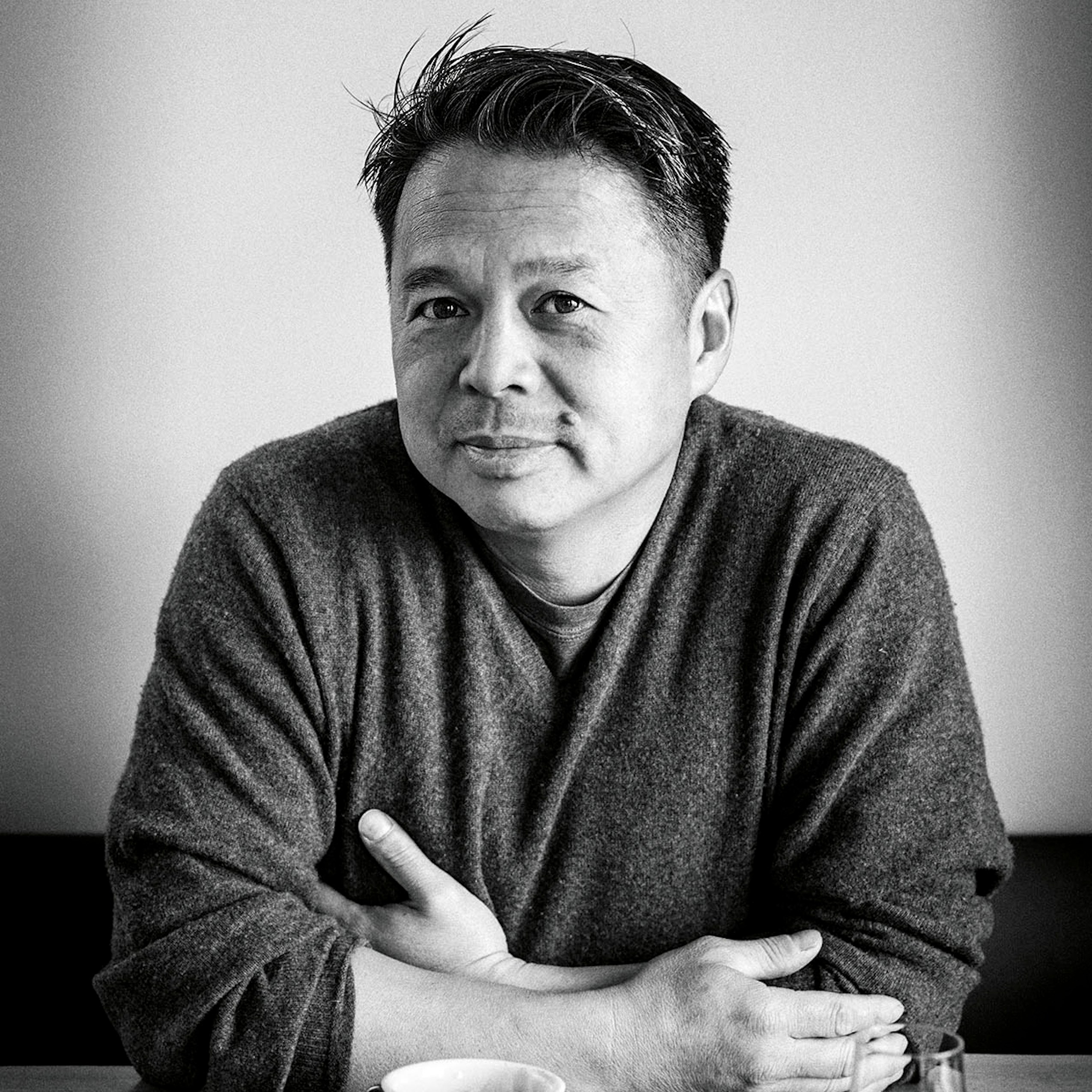A man wearing a dark sweater sits at a table with a mug of coffee.