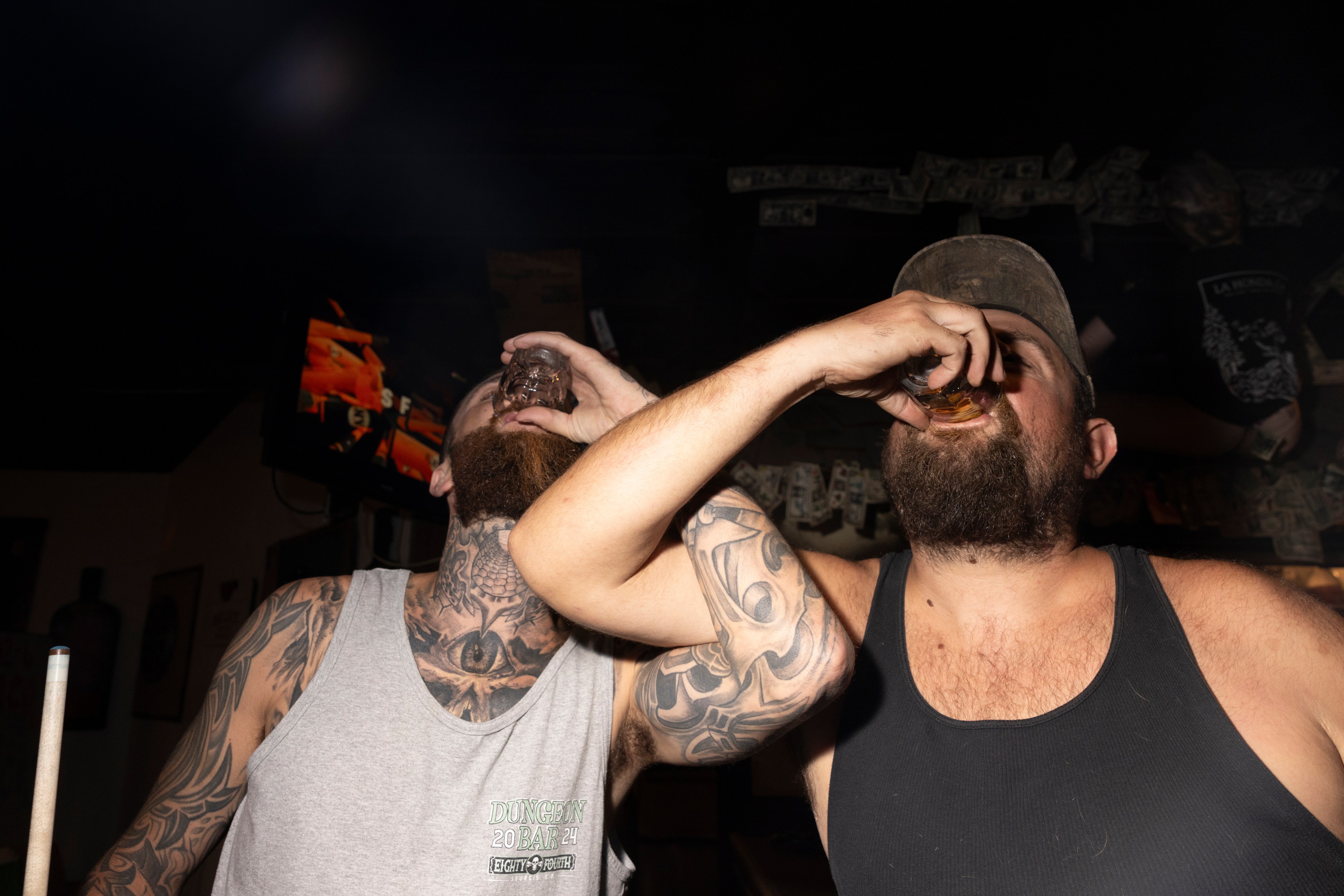 Two men with tattoos and beards are taking shots in a dimly lit bar, with dollar bills on the walls and a TV screen showing a fiery image in the background.