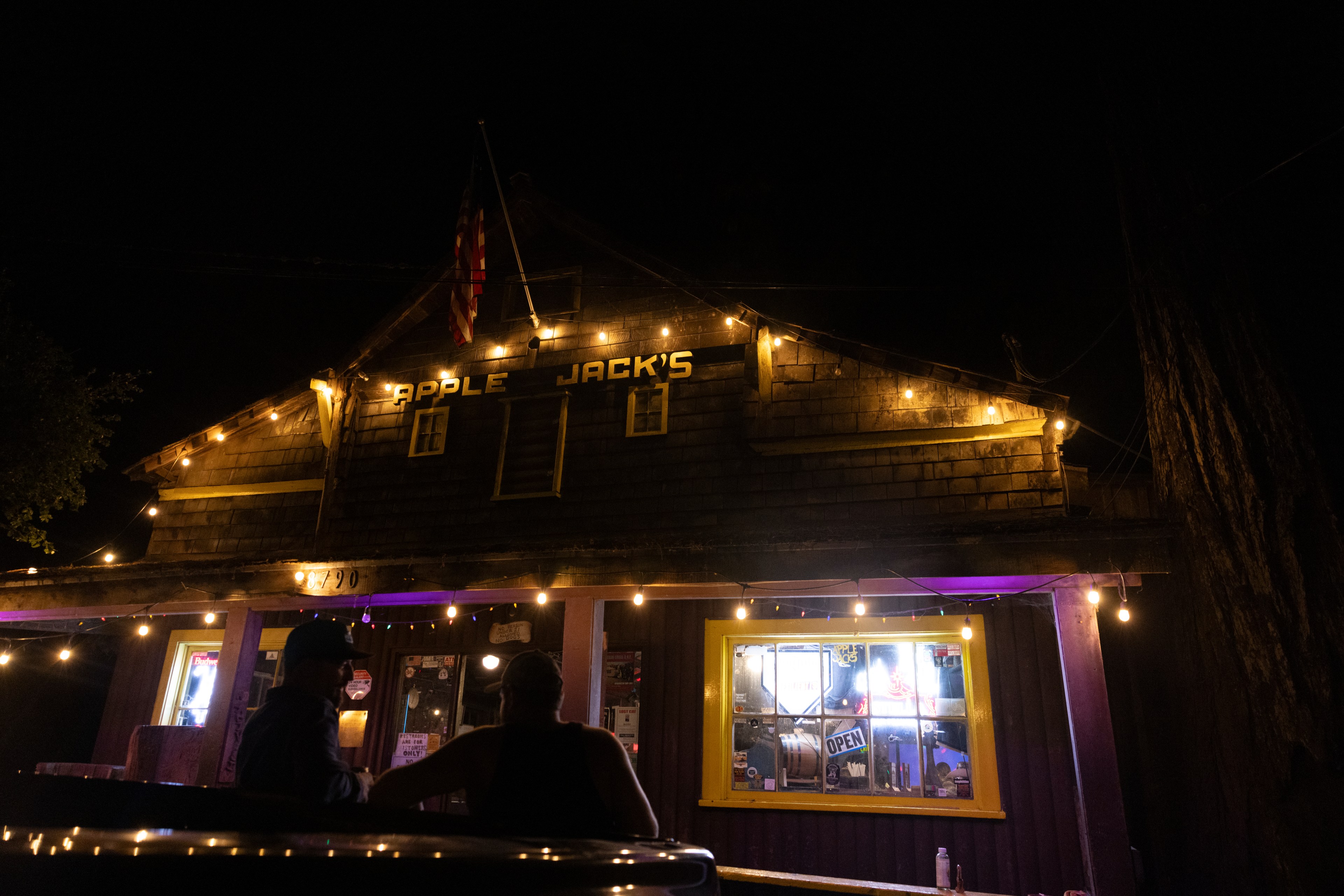 The image shows a dimly lit rustic building named &quot;Apple Jack's&quot; at night, decorated with string lights and an American flag, with two people seated outside.