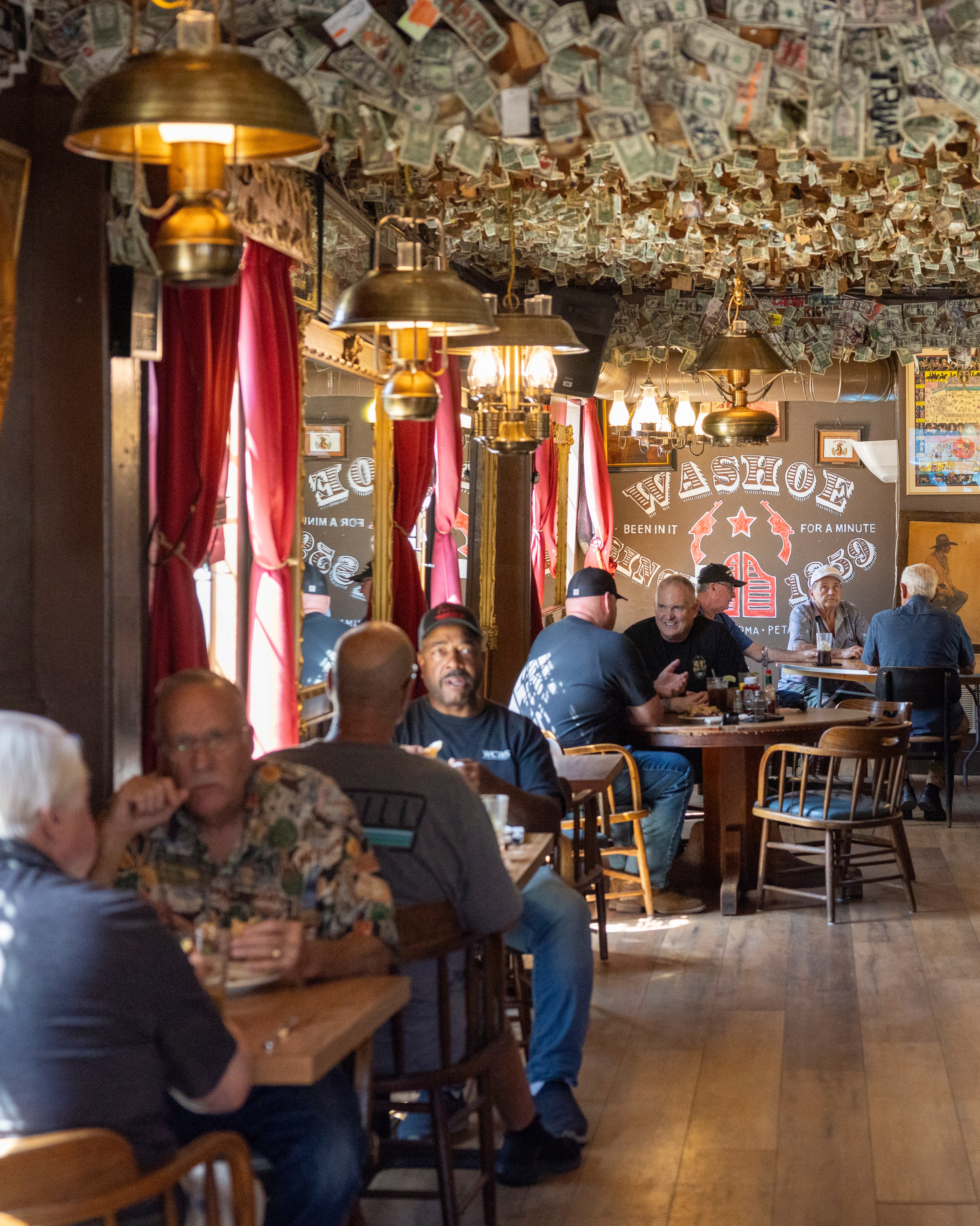 Patrons eat at a rustic restaurant with dollar bills covering the ceiling. Red curtains and warm pendant lights add to the cozy ambiance.