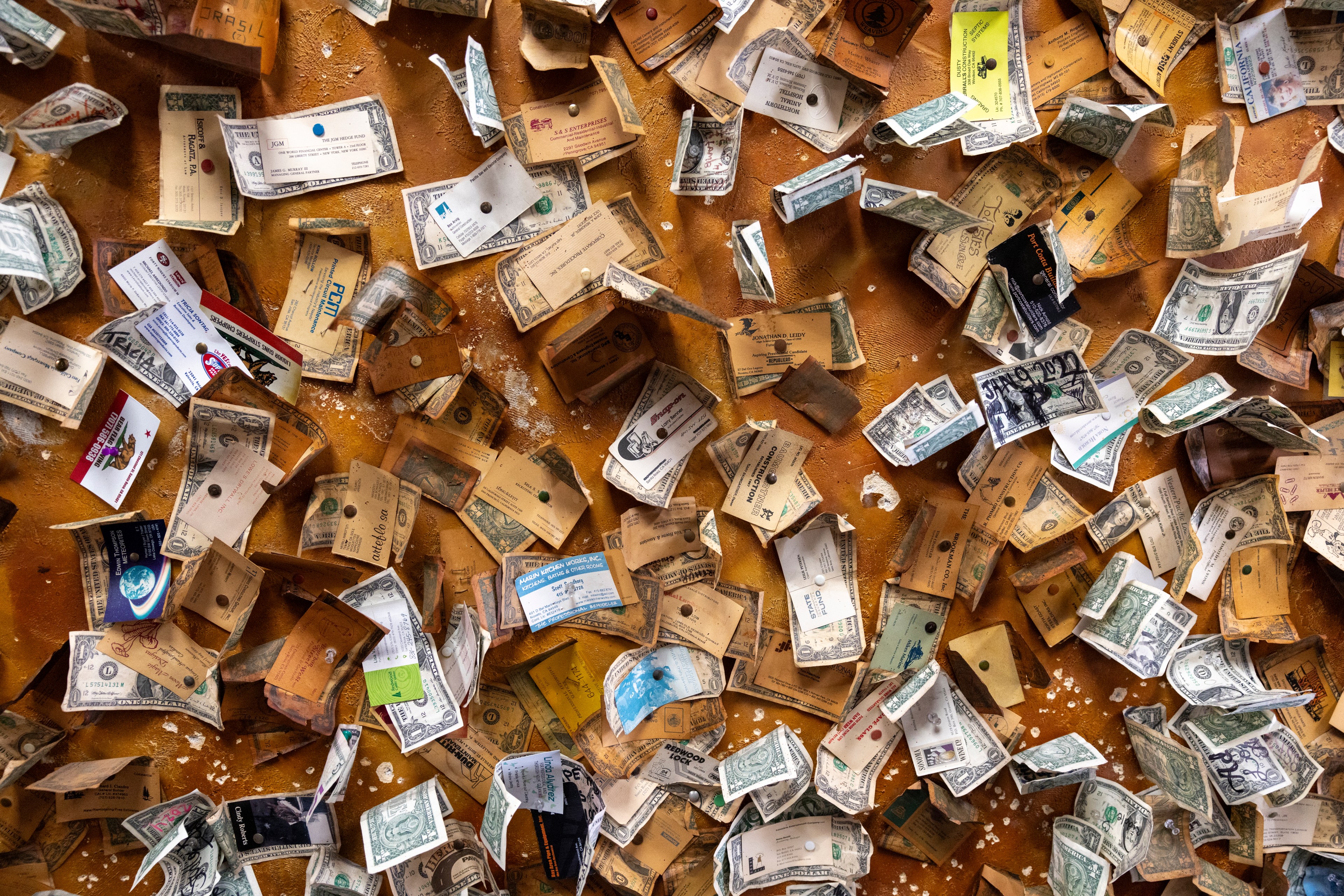 The image shows numerous business cards, dollar bills, and other pieces of paper pinned to an orange corkboard in a cluttered, overlapping manner.