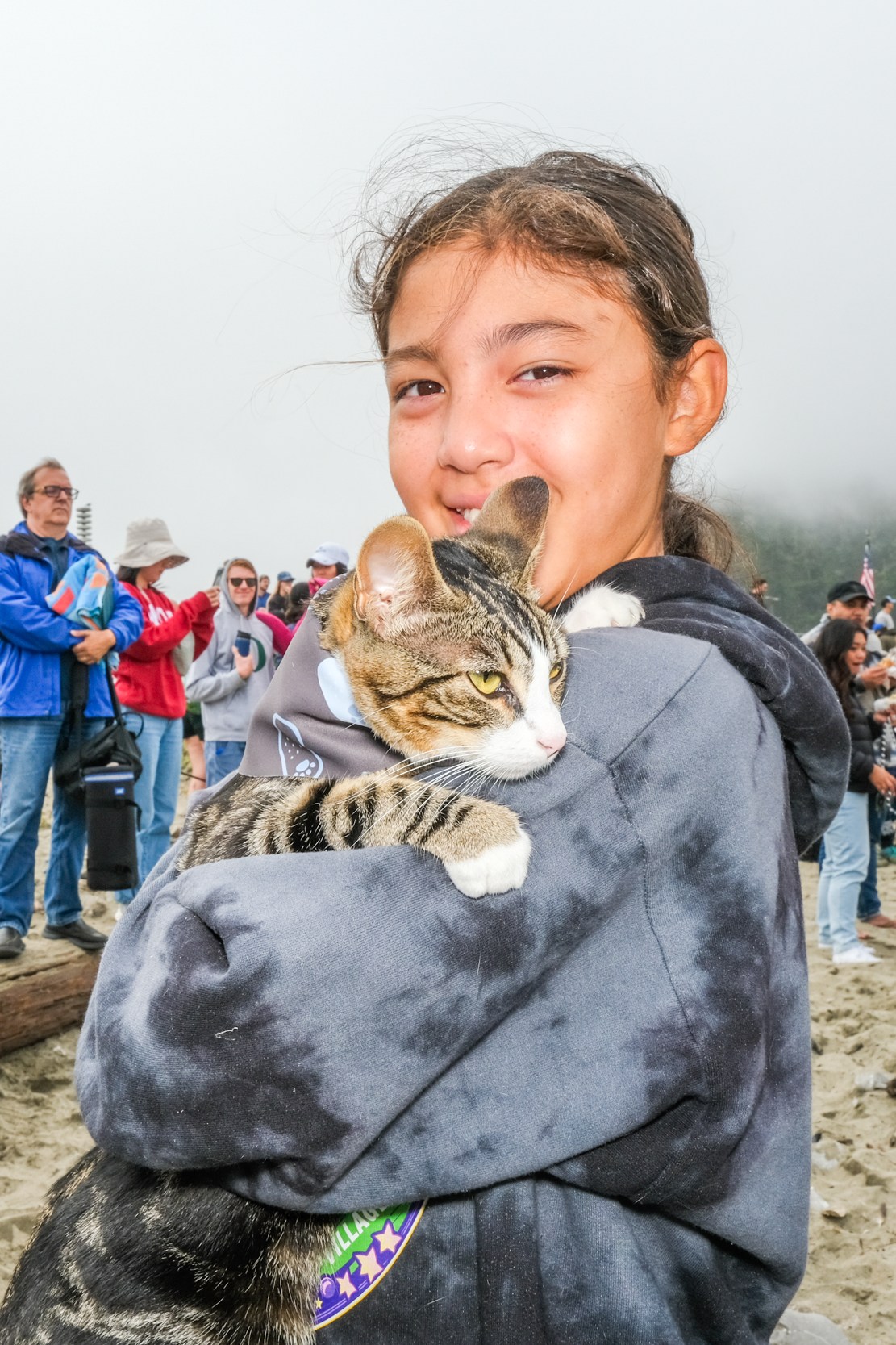 A girl is holding a cat tightly, smiling at the camera, while a group of people behind her are watching and taking photos on a foggy beach.