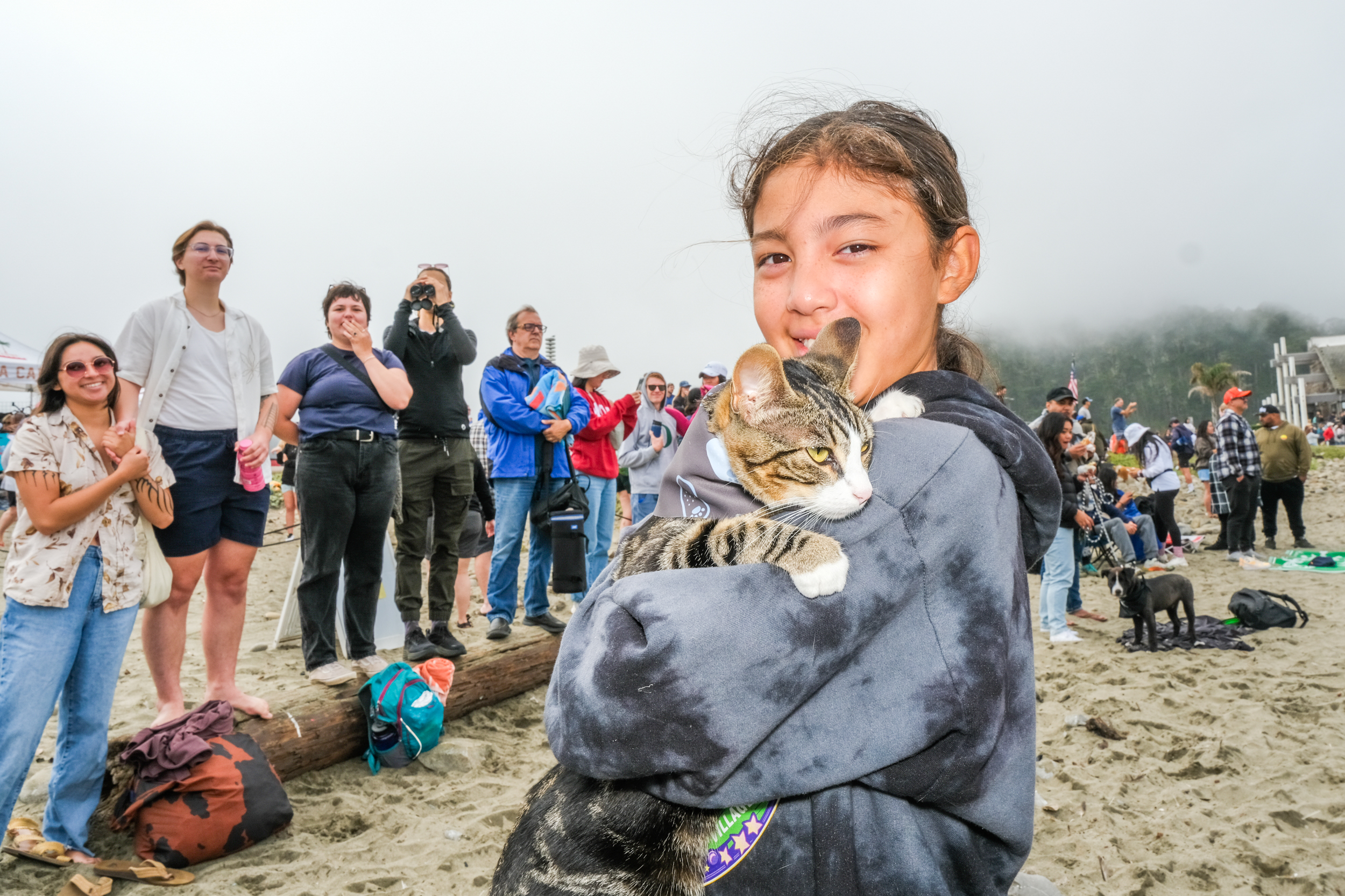 A girl is holding a cat tightly, smiling at the camera, while a group of people behind her are watching and taking photos on a foggy beach.