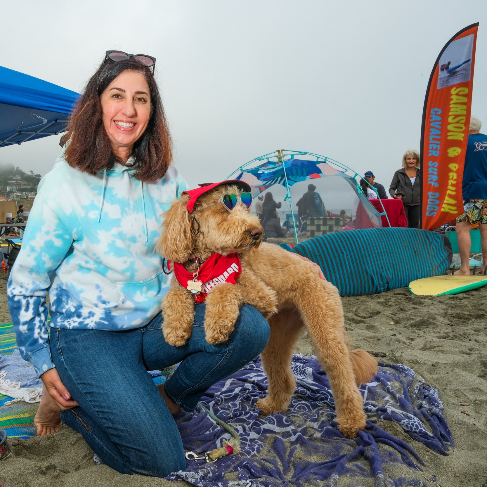 A smiling woman in a blue tie-dye hoodie kneels on a beach next to a dog wearing sunglasses and a red bandana. Beachgoers and tents are in the background.