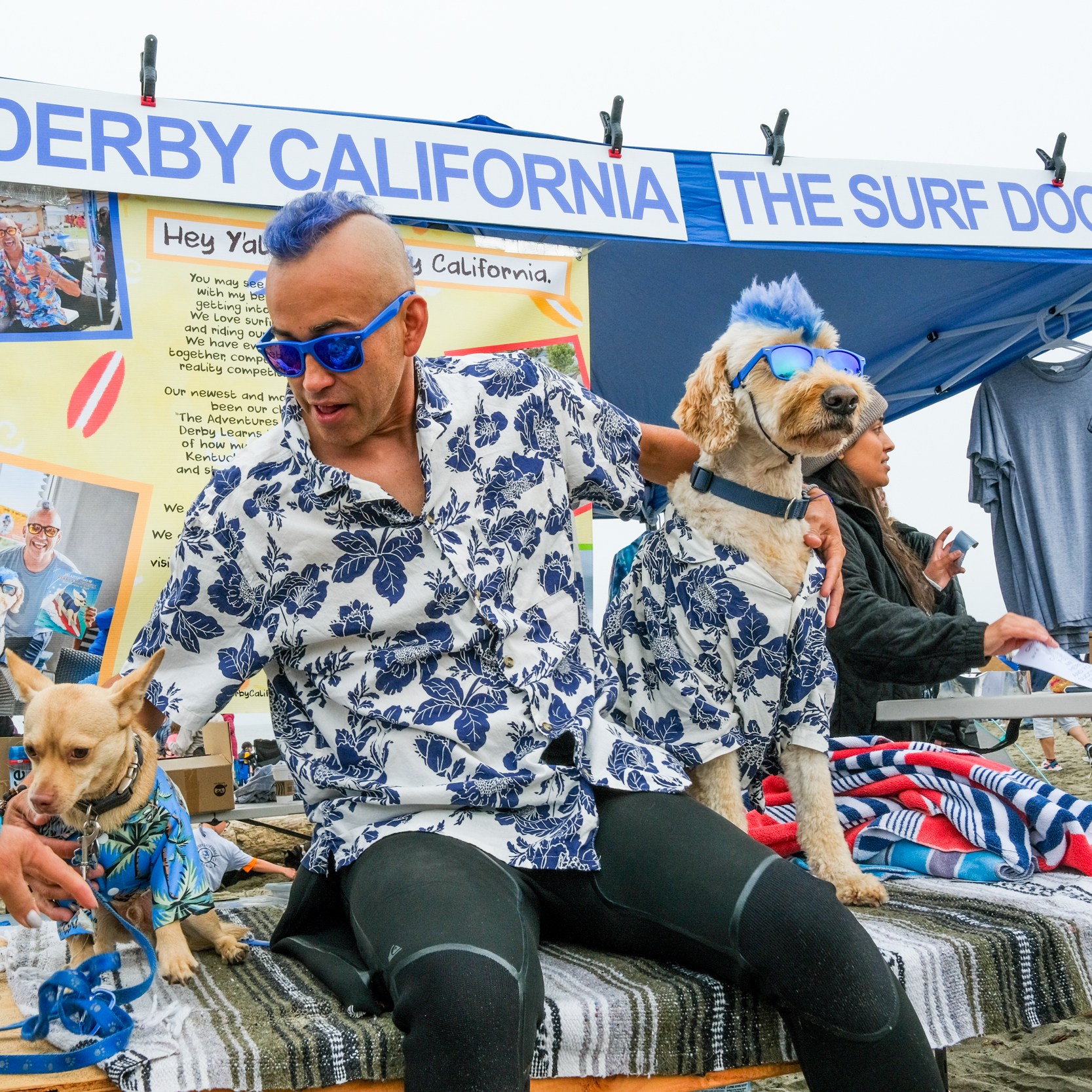 A man and two dogs, all wearing matching Hawaiian shirts, sit at a surf dog booth. One dog also wears sunglasses, while someone interacts with the smaller dog.