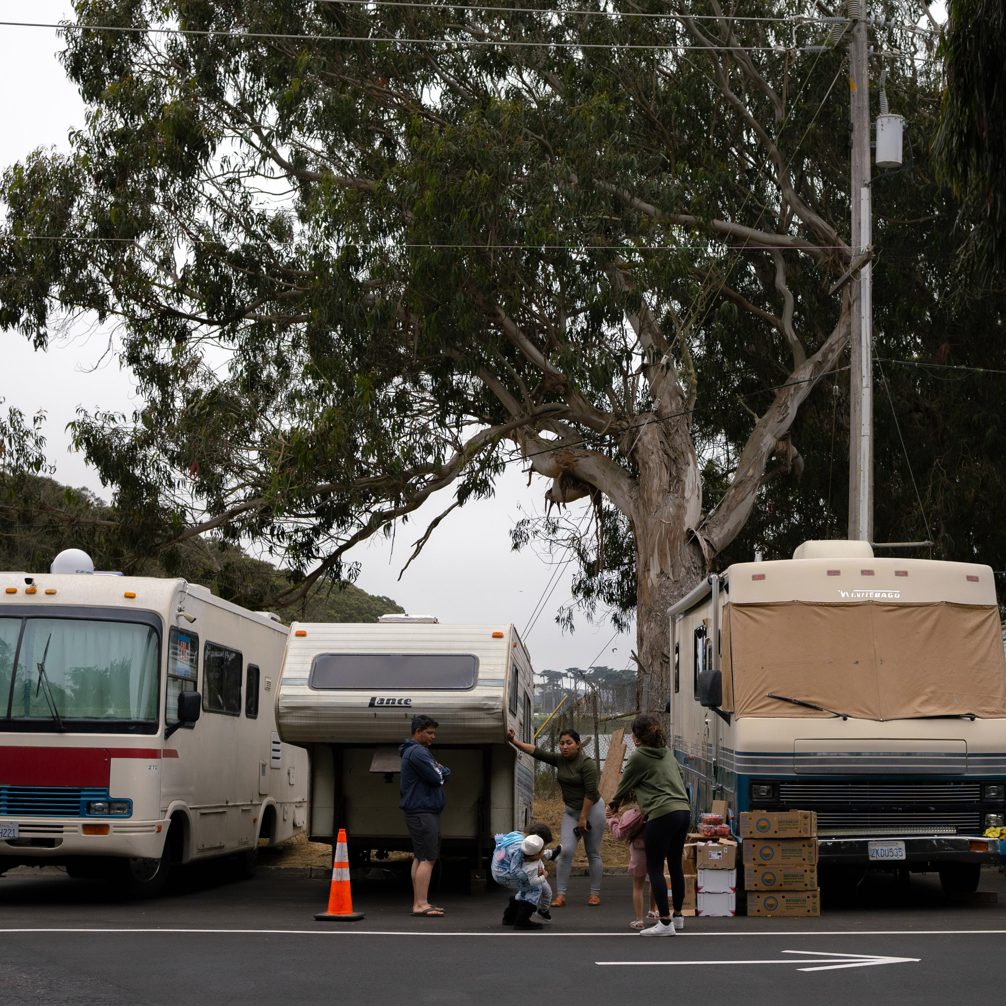 The image shows two RVs and a camper parked near a tree. Four people interact in front, with one person squatting and three standing; boxes and a traffic cone are nearby.