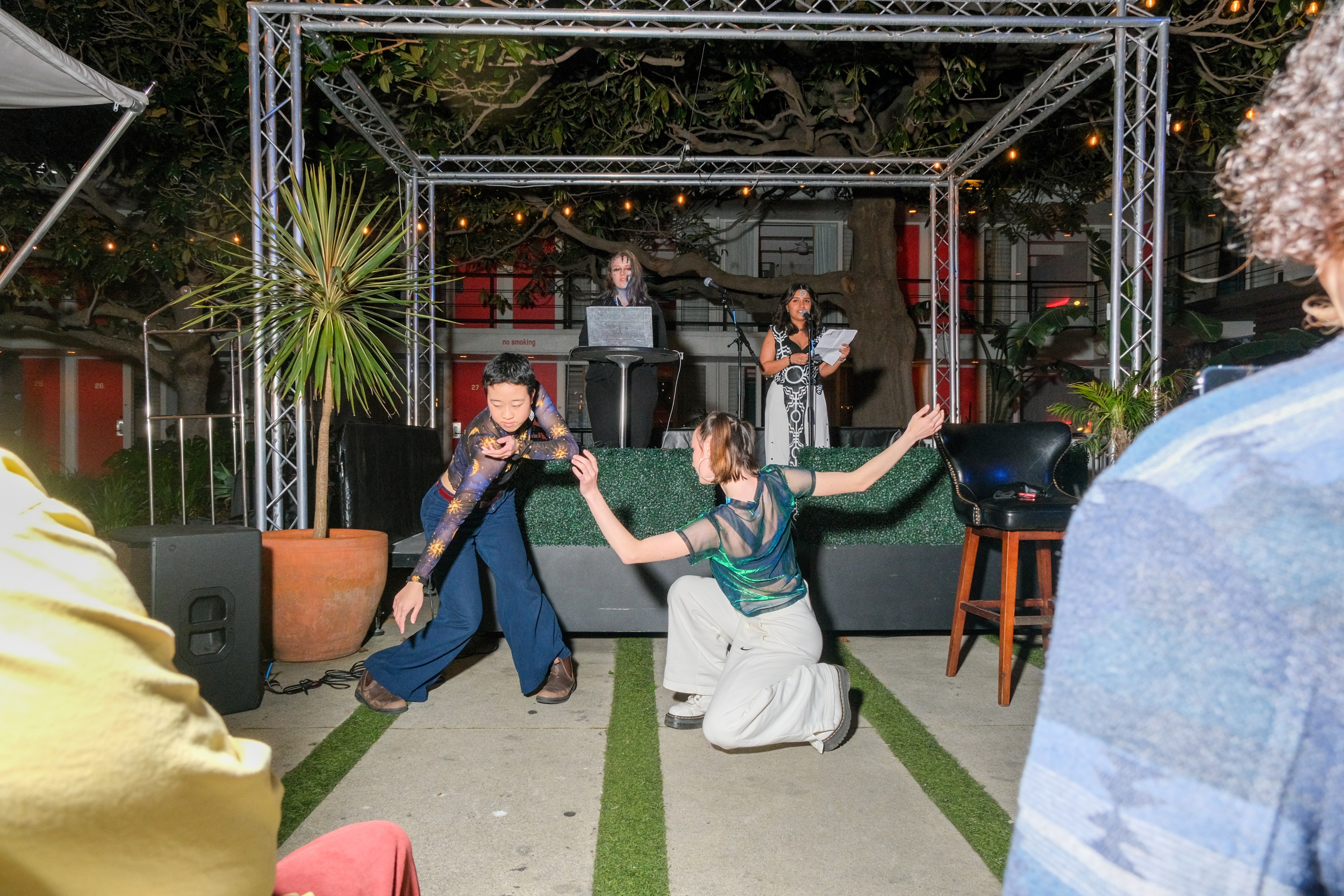 Two dancers perform on the floor in front of a small stage with two people, one at a podium and one with a microphone, while onlookers watch.