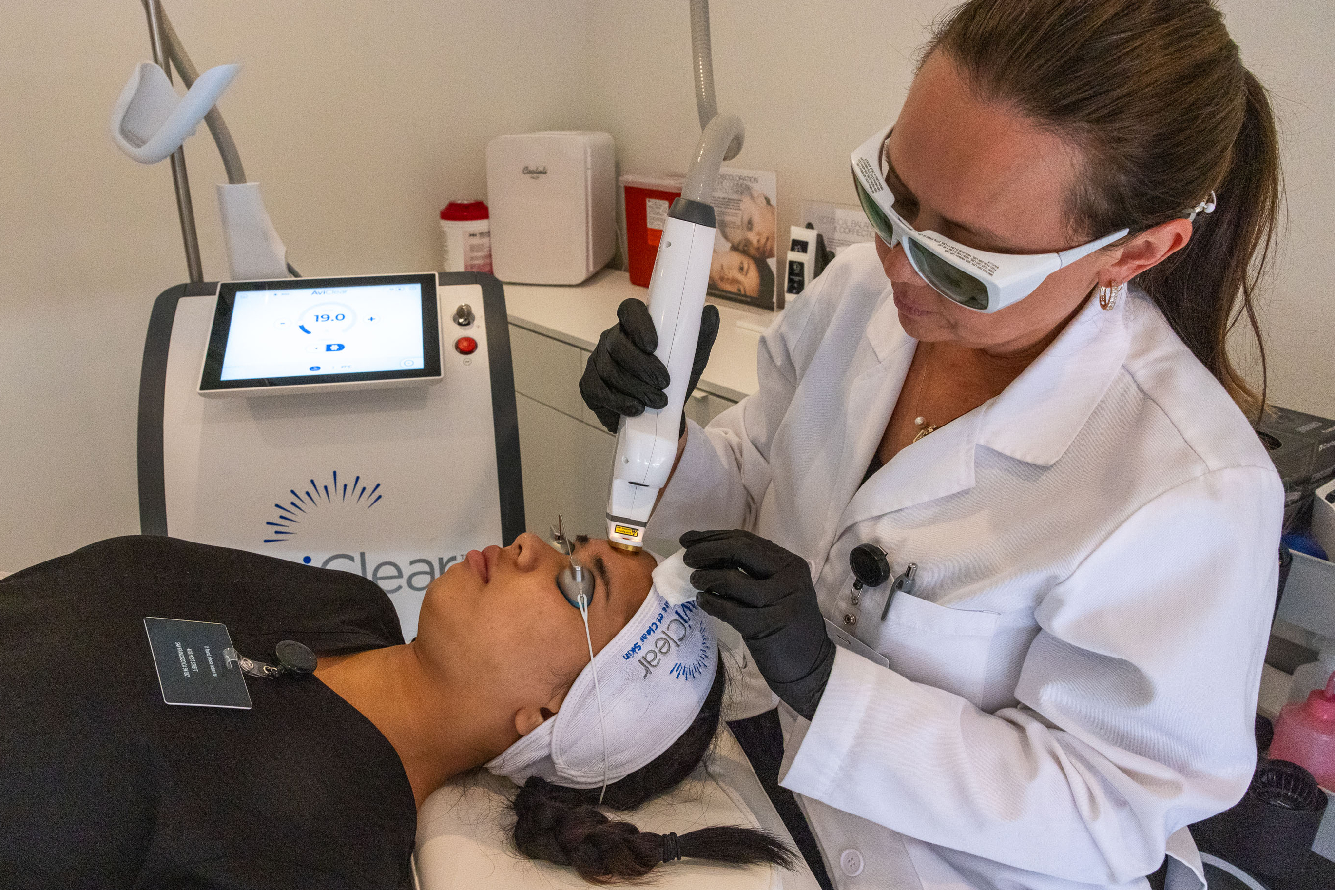 A woman in a white lab coat and protective glasses performs a facial treatment on a reclining person using a handheld device, with a screen displaying &quot;19.0&quot;.