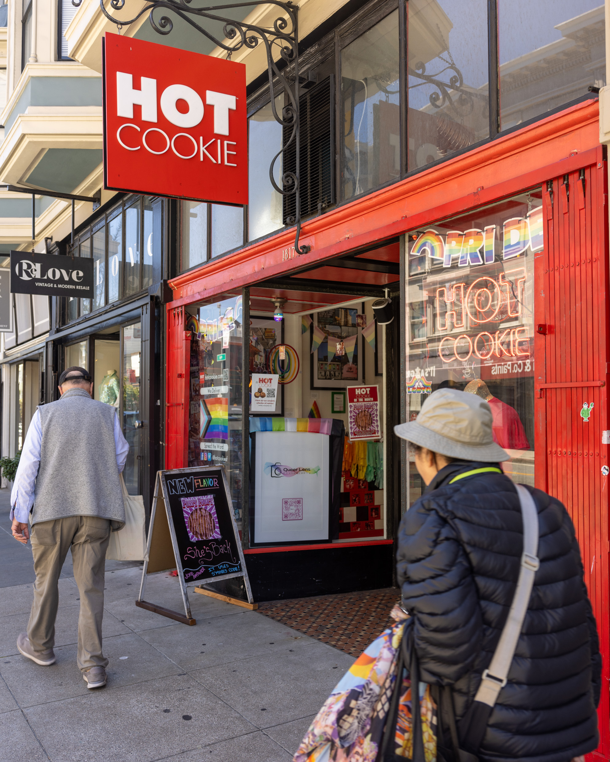 Two people walk past a brightly colored store with a red &quot;HOT COOKIE&quot; sign. The storefront has a chalkboard advertising a new flavor, and colorful decorations inside.