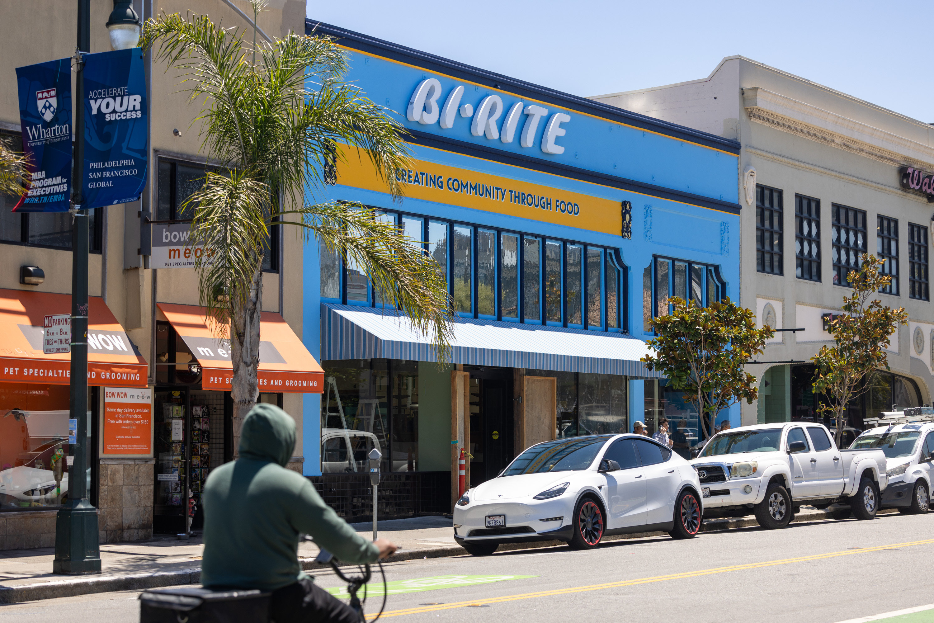 A cyclist rides past a blue building with &quot;Bi-Rite&quot; written on it. Parked vehicles, including a white Tesla, line the street. Nearby is an orange-awning pet store.