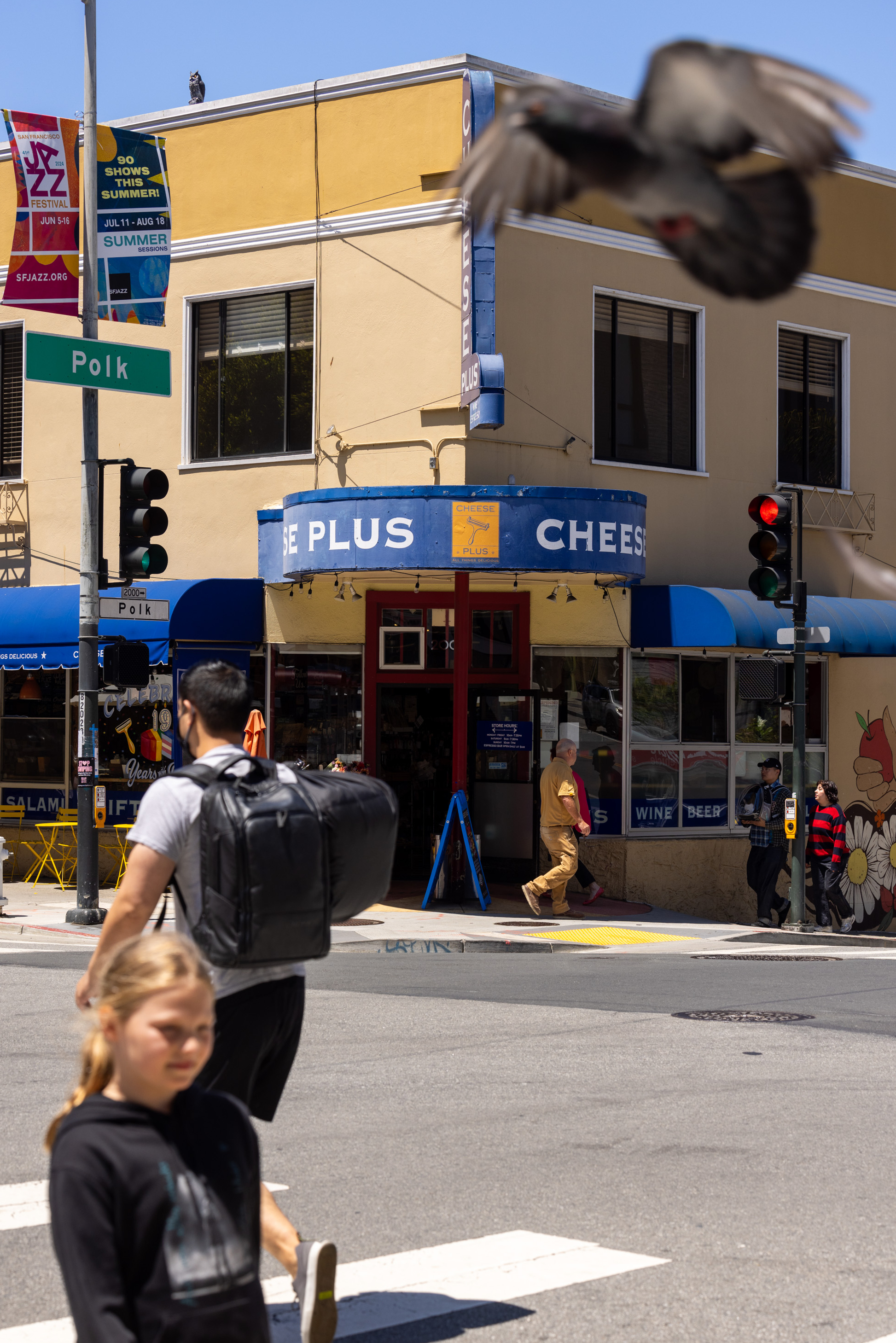 A corner street view shows a yellow building with &quot;Cheese Plus&quot; signage. People cross the street, and a blurred pigeon flies by. A green Polk street sign is visible.