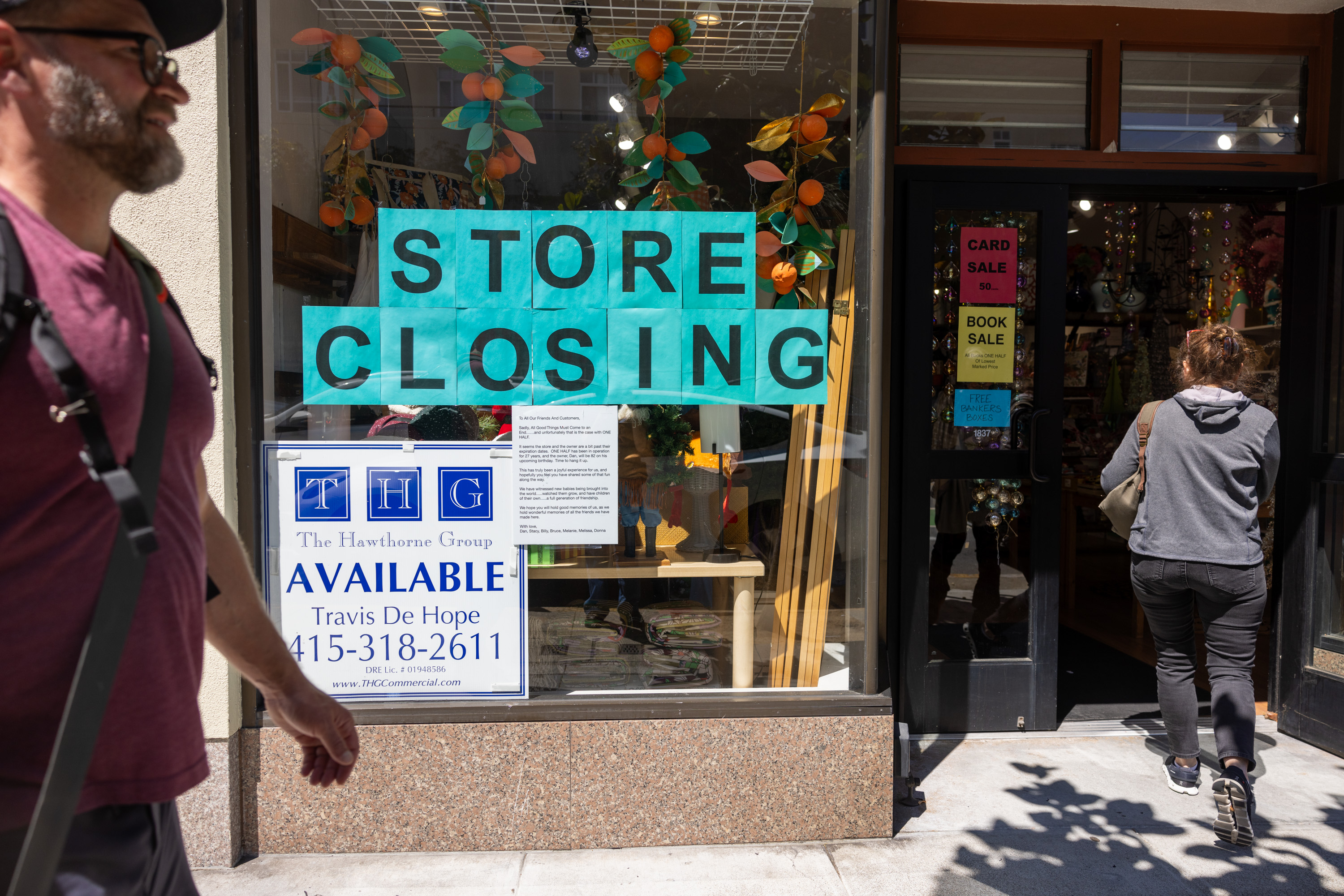 A storefront with signs reading &quot;STORE CLOSING&quot; and &quot;AVAILABLE&quot; with contact details. One person walks past, while another enters. Decorations are visible inside.