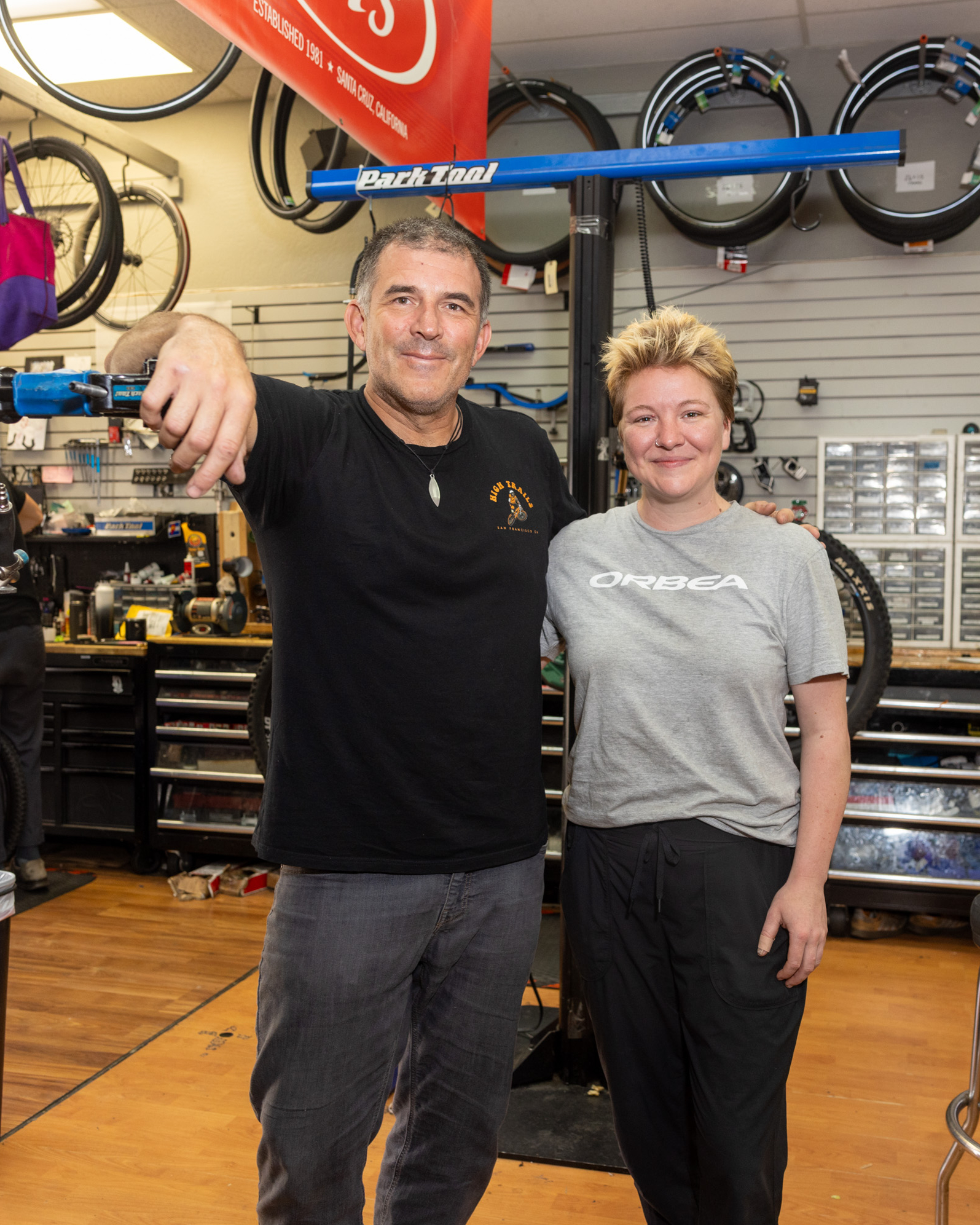 Two smiling individuals are standing inside a bike shop. The person on the left leans on a blue bike repair stand, and the person on the right has their hand on the other's back.