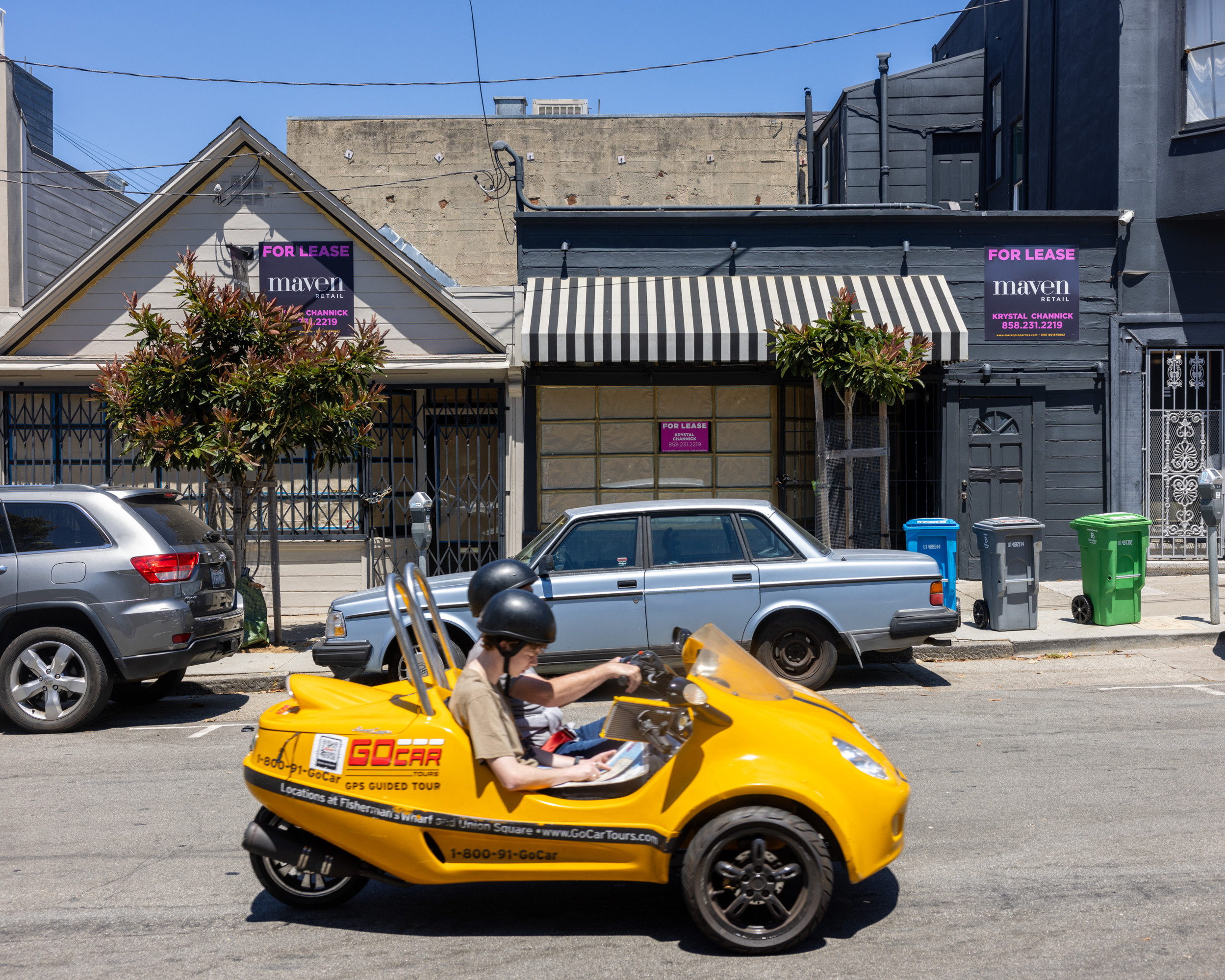 A yellow GoCar with two helmeted passengers drives past parked cars and two "For Lease" signs on a street with gray buildings and a striped awning.