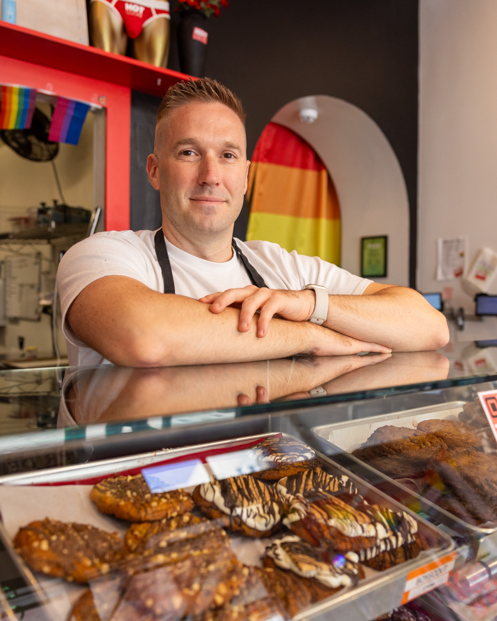 A person wearing a white shirt and an apron stands behind a glass display case filled with various pastries. A rainbow flag and colorful decor are visible in the background.