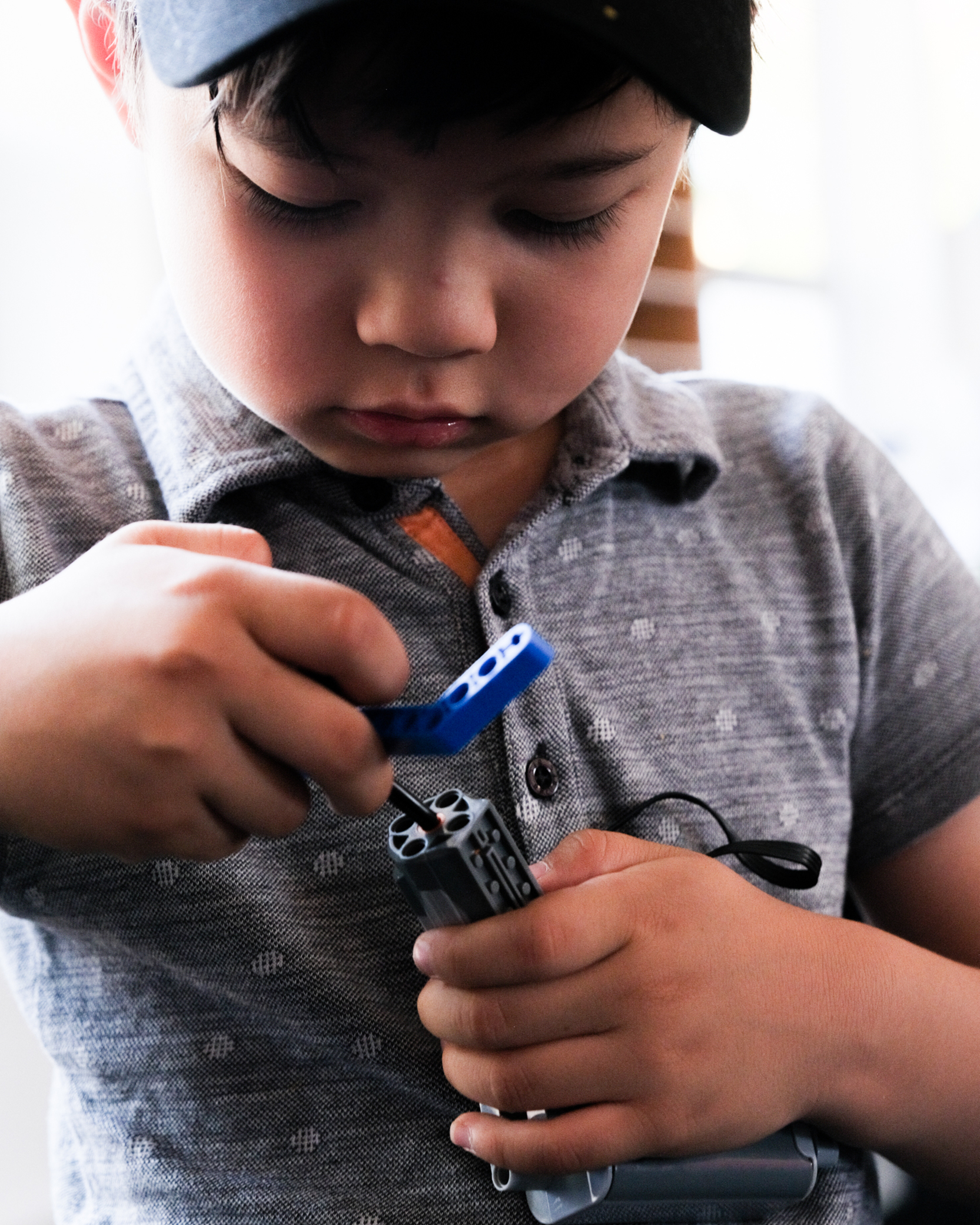 A young child wearing a black cap and a grey shirt intently focuses on assembling a blue and grey Lego-like toy with both hands.