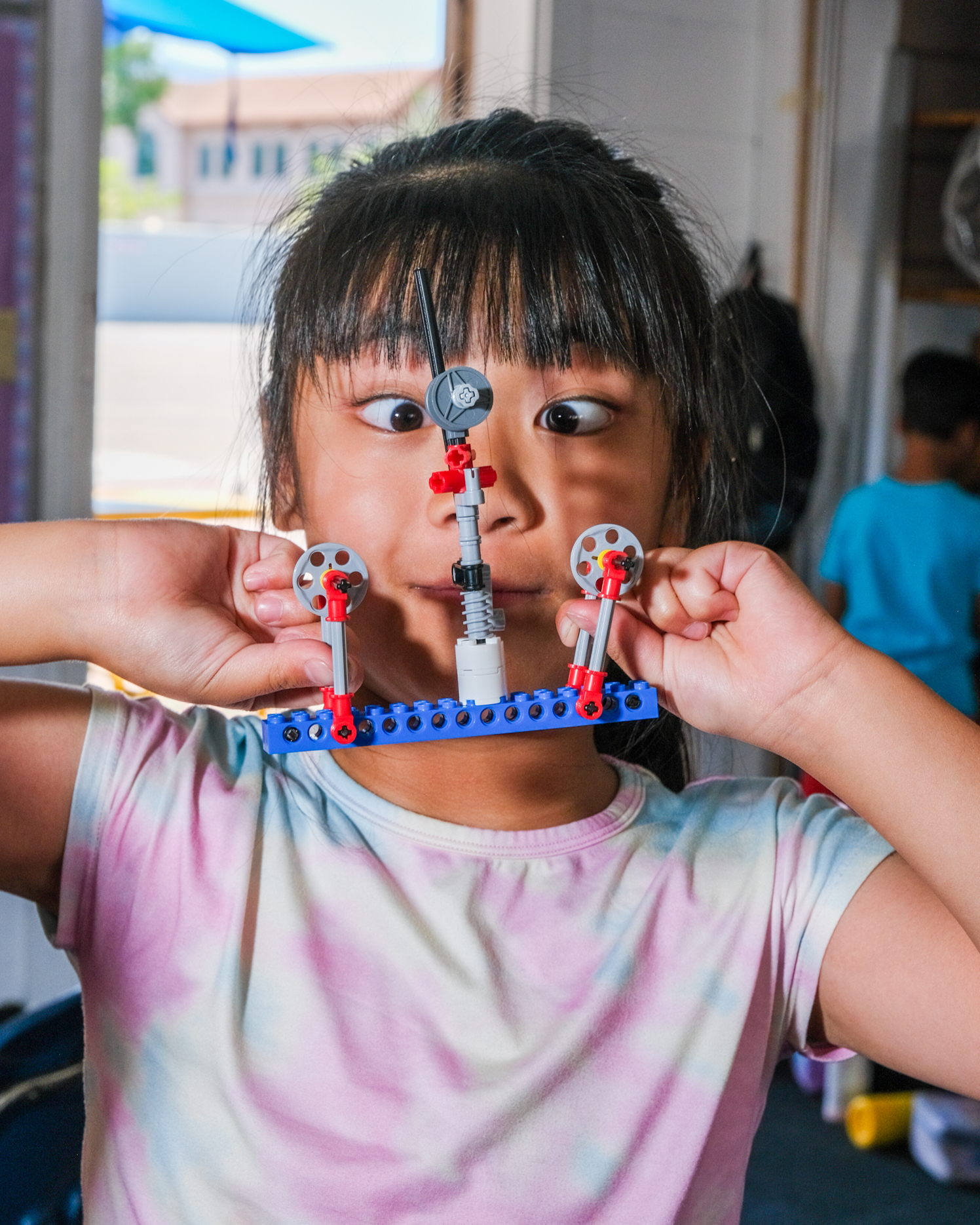 A girl with crossed eyes holds a colorful LEGO creation in front of her face while making a funny expression in a playful indoor setting.