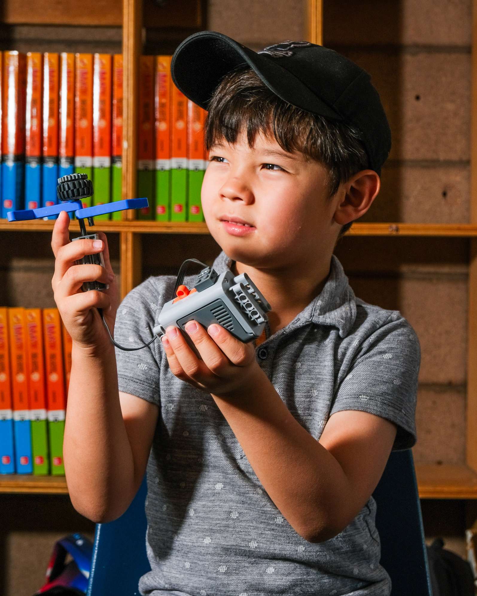 A young boy in a black cap and gray shirt intently examines a mechanical toy in his hands, with books arranged in shelves behind him.