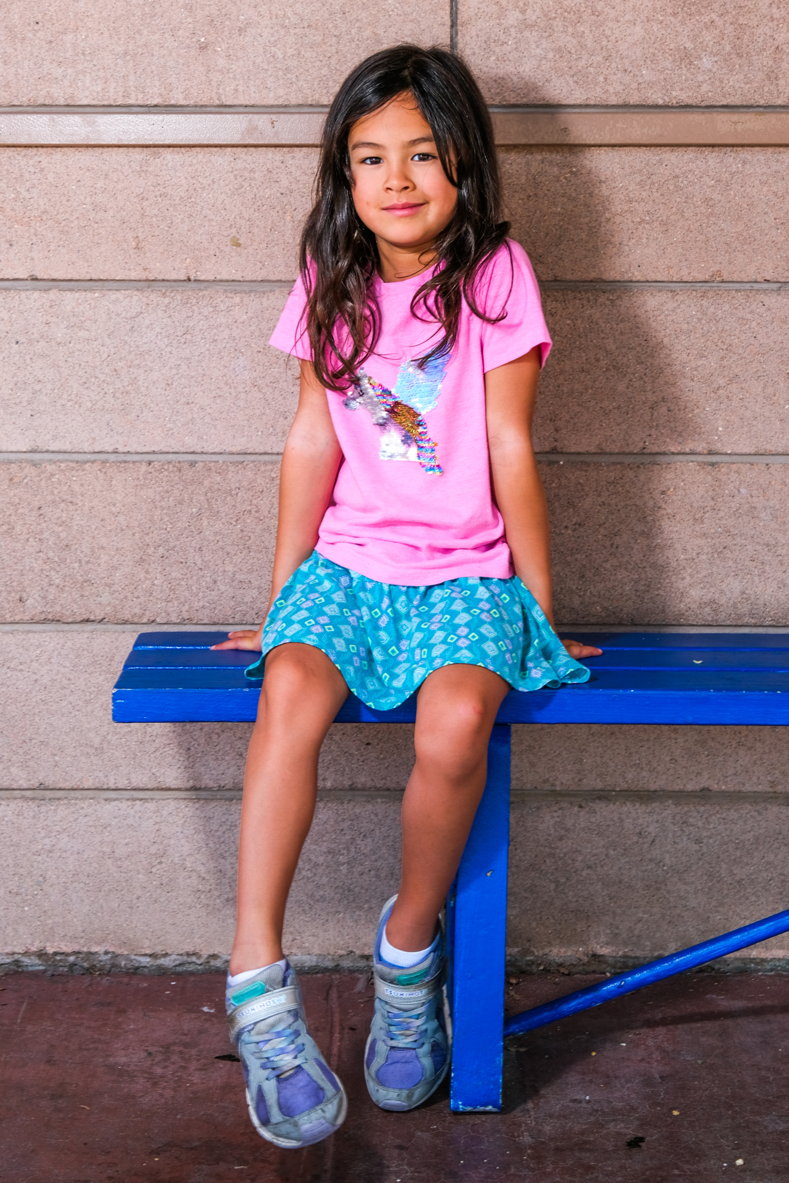 A young girl with long dark hair, wearing a pink T-shirt with a sequin design and a blue patterned skirt, sits on a blue bench against a beige brick wall.