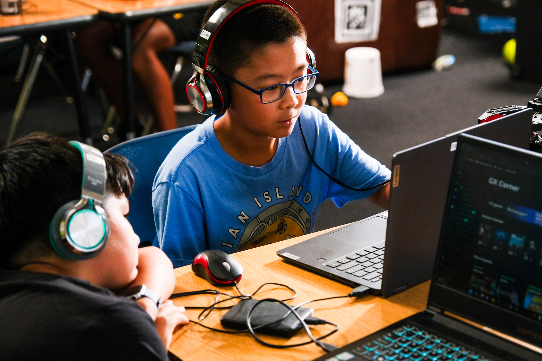 Two boys with headphones sit at a table using laptops; one is actively working, while the other watches intently.