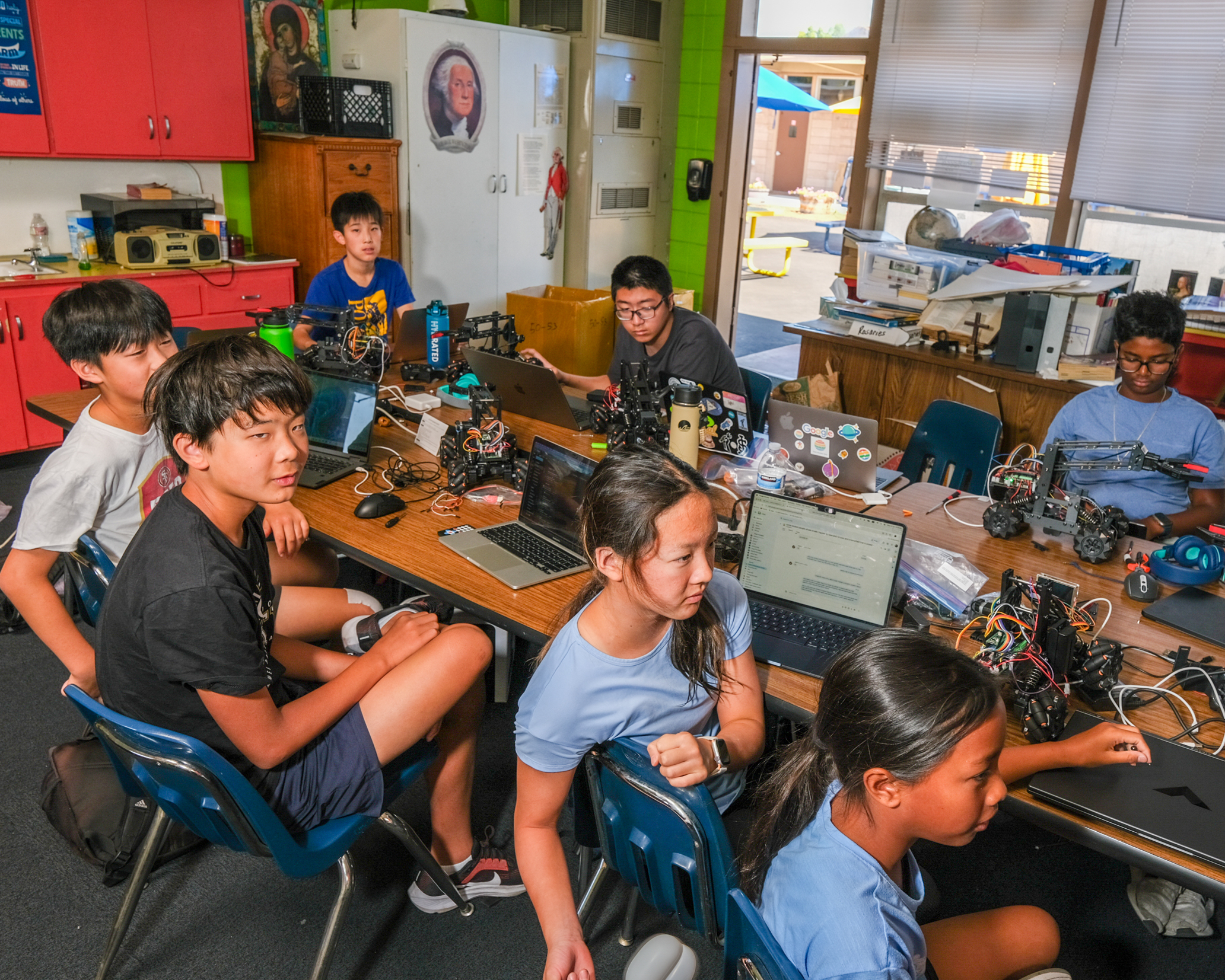 A group of eight young people are sitting around a table with laptops, robots, and other tech equipment, actively working and collaborating in a classroom setting.