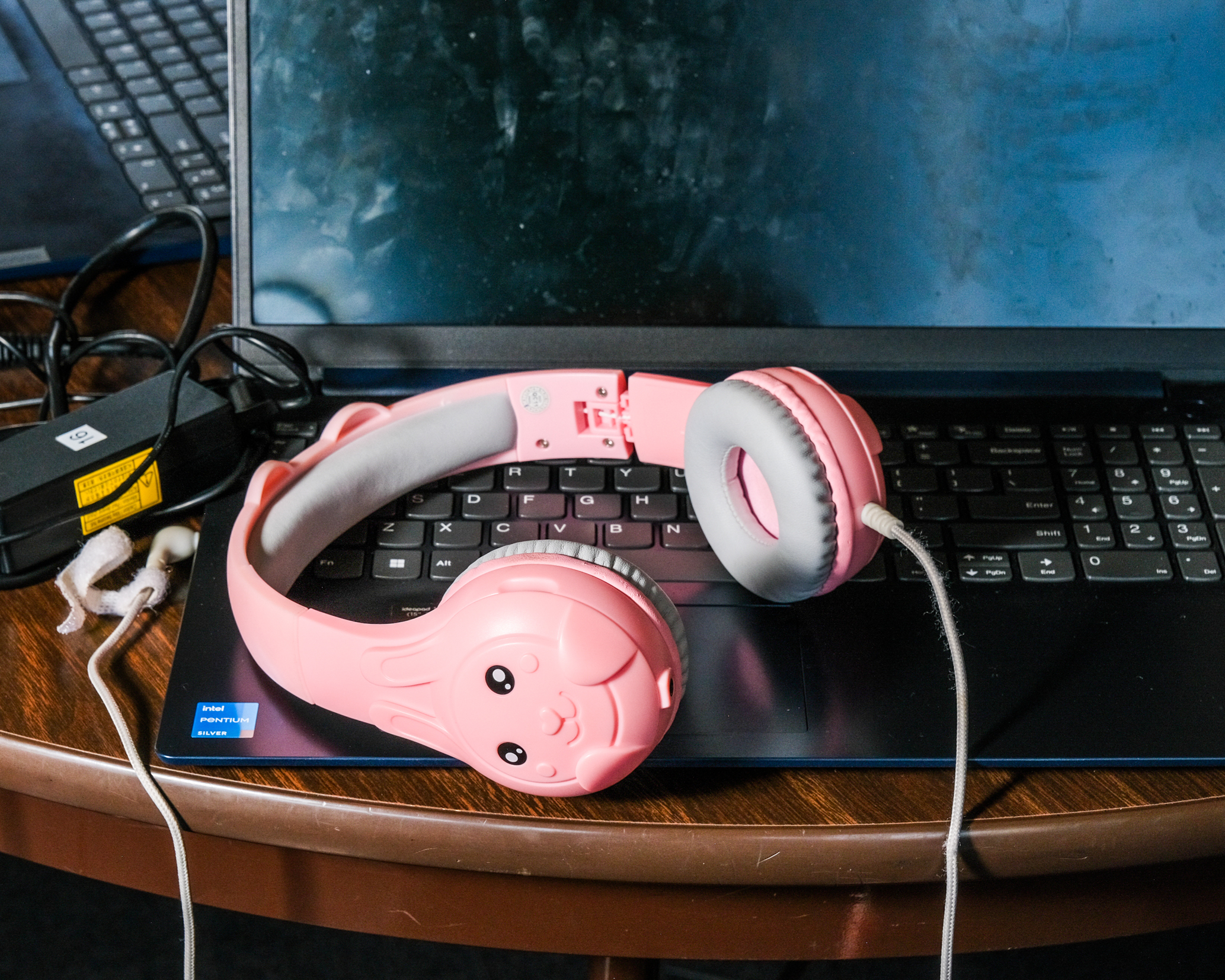 A pair of pink headphones with a cute animal face design rests on a laptop's keyboard, which has a slightly smudged screen. The laptop charger is visible beside it.