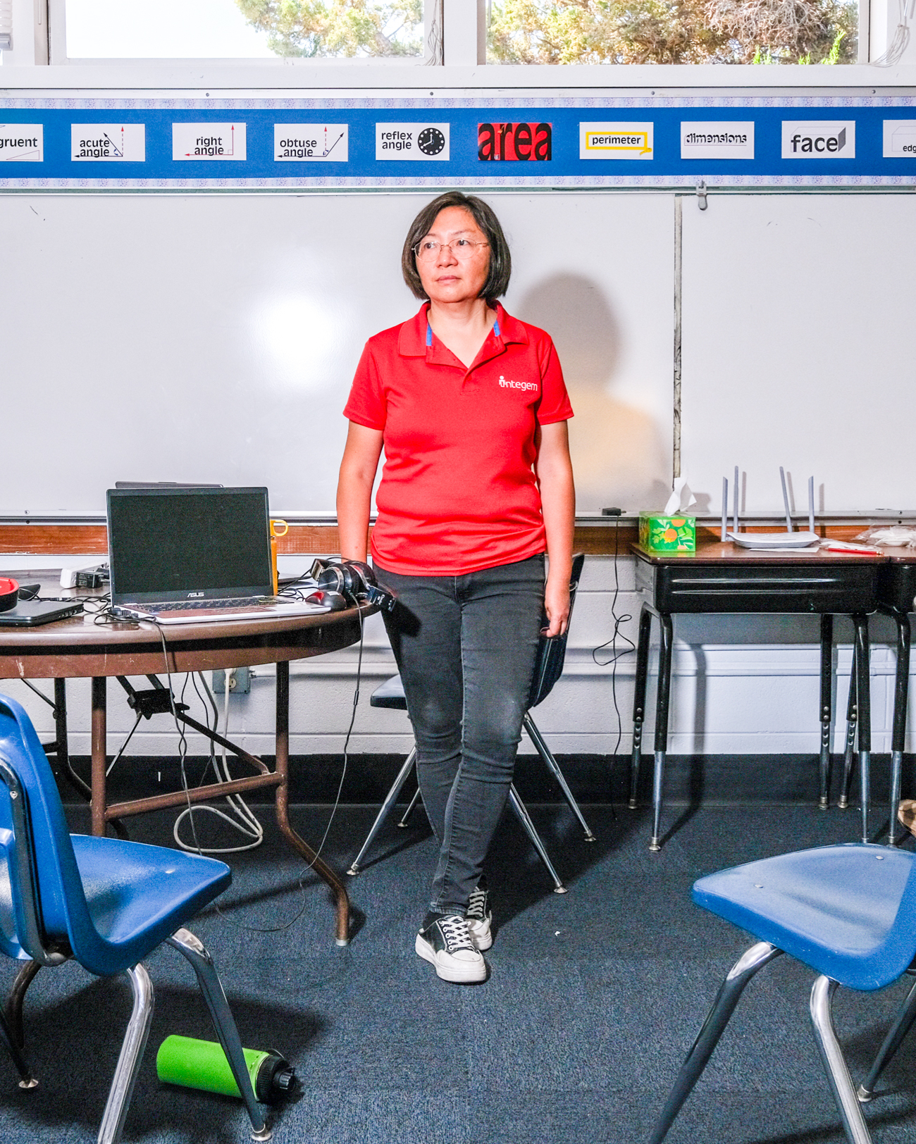 A person wearing a red &quot;Integem&quot; shirt stands in a classroom, with a laptop and other devices on a table. Math terms are displayed on a board behind them.