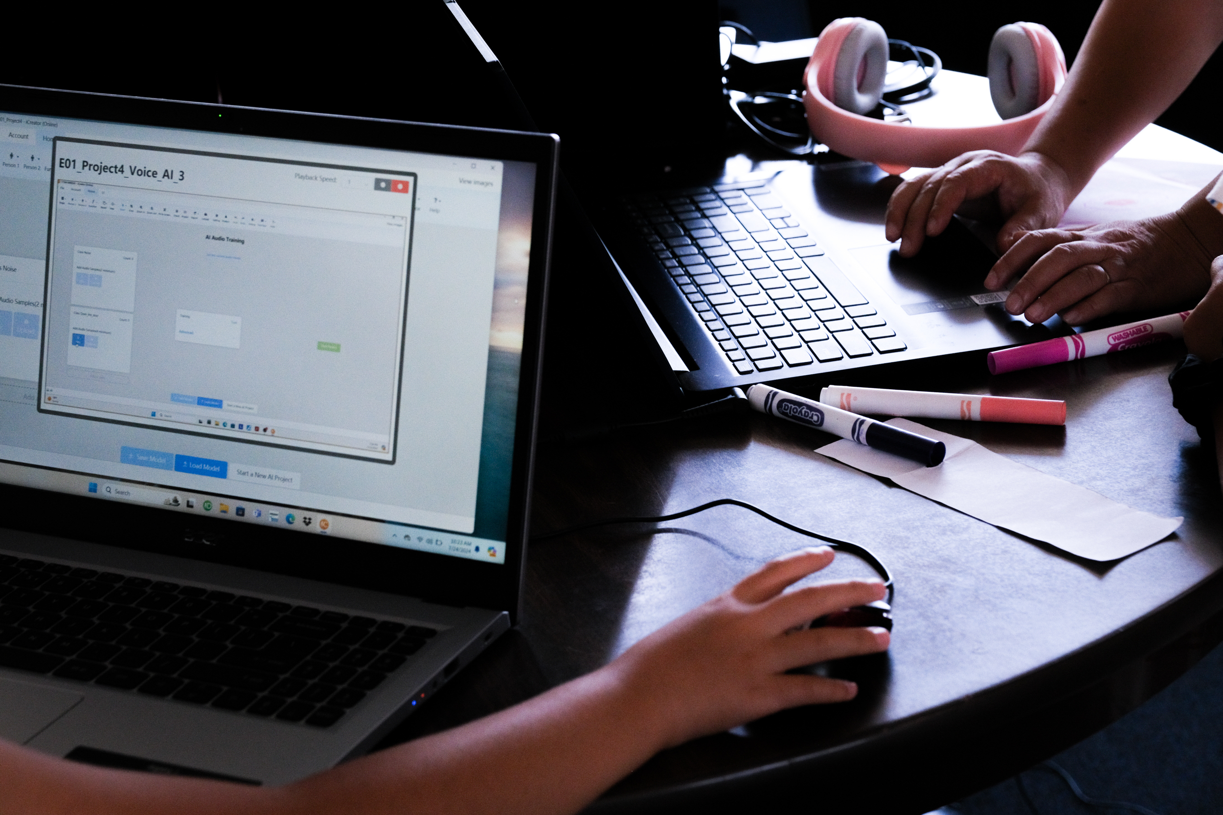 Two people are working on laptops at a table with pink headphones and various markers. One screen displays a project titled &quot;E01 Project4 Voice AI_3.&quot;