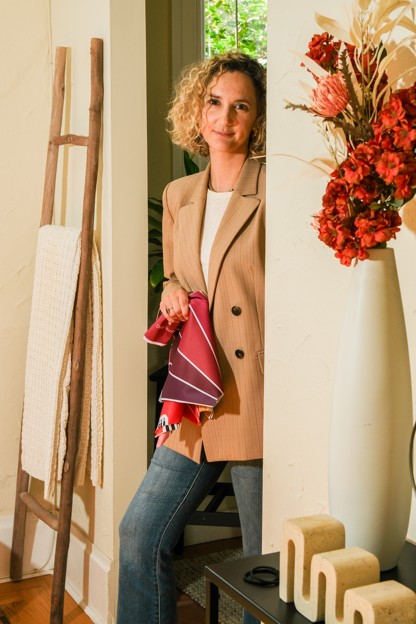 A woman with curly hair leans against a doorway wearing a beige blazer and holding a colorful scarf. Beside her, a ladder holds a knitted blanket, and a vase with red flowers is on a table.