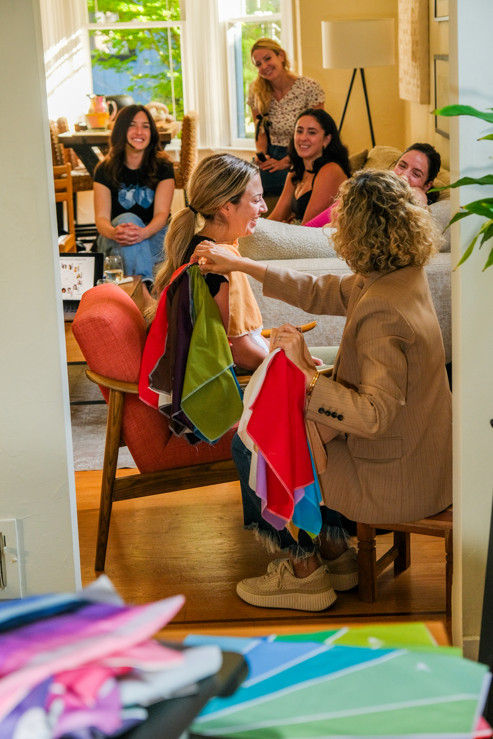 A group of six women enjoys a colorful fabric styling session in a cozy, sunlit living room. One woman is choosing fabrics for another, while the others watch and laugh jovially.