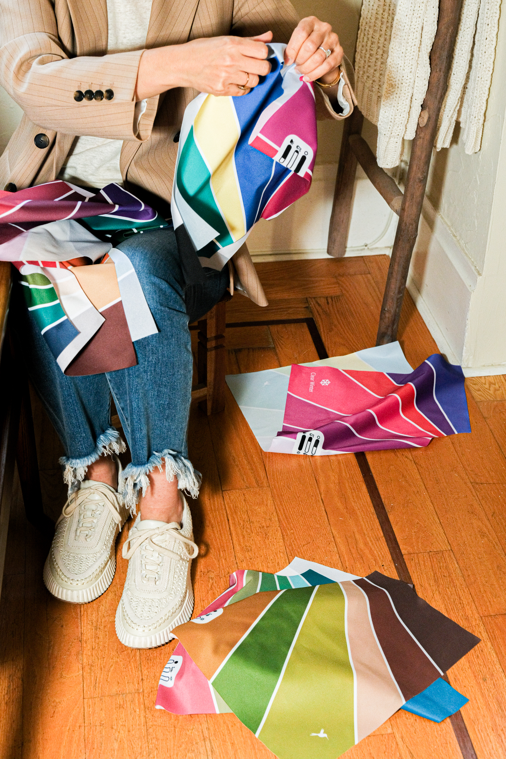 A seated individual wearing a beige blazer and frayed jeans handles vibrant fabric swatches, some of which are on their lap and scattered on a wooden floor.