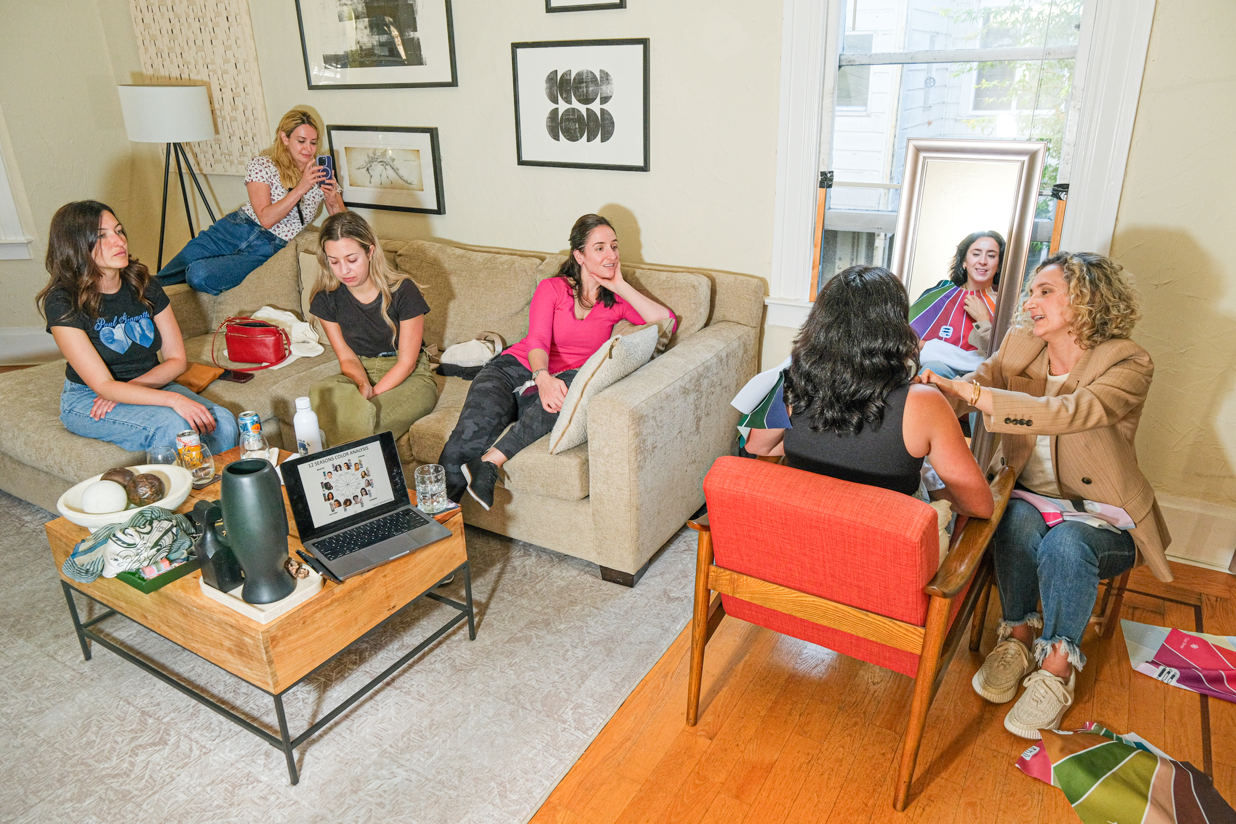 Six women are in a cozy living room. Some are chatting, while one takes photos and another helps another woman with colorful fabrics by a large mirror.