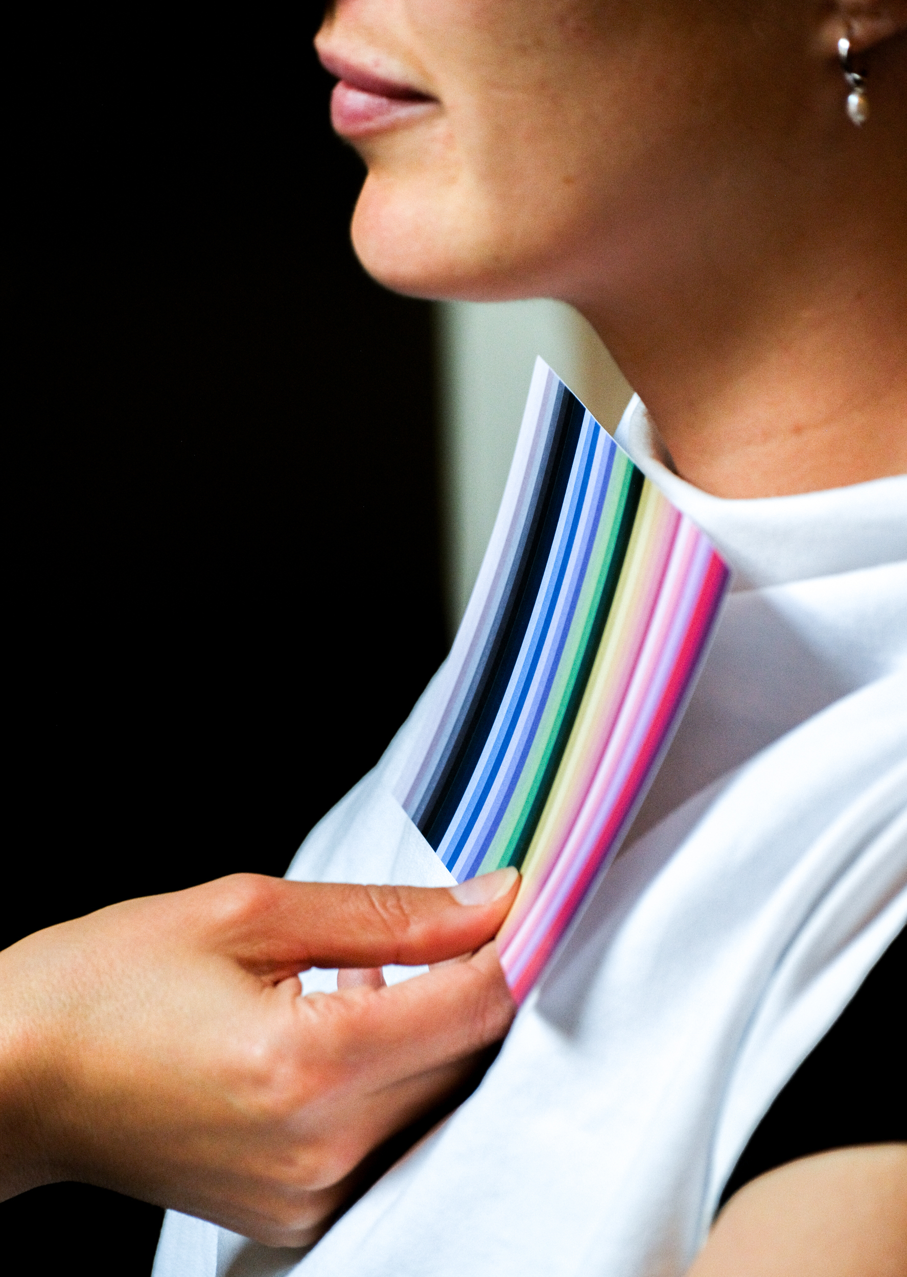 A person holds a fan of multicolored paper strips near their neck, wearing a white shirt and an earring. The background is dark and out of focus.