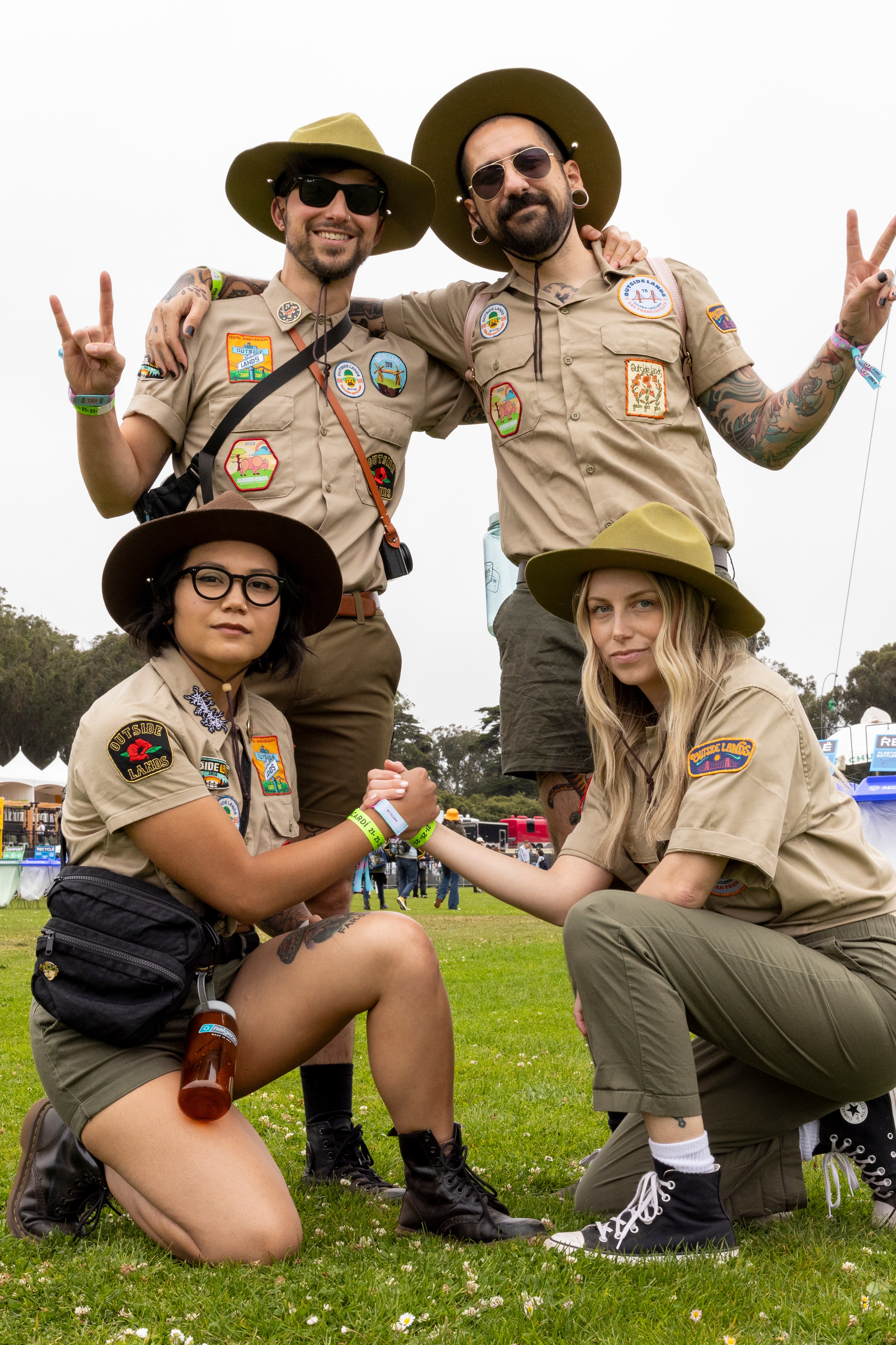 Four people in scout uniforms, two standing and two kneeling, pose on a grassy field at what appears to be an outdoor event with tents and a stage in the background.