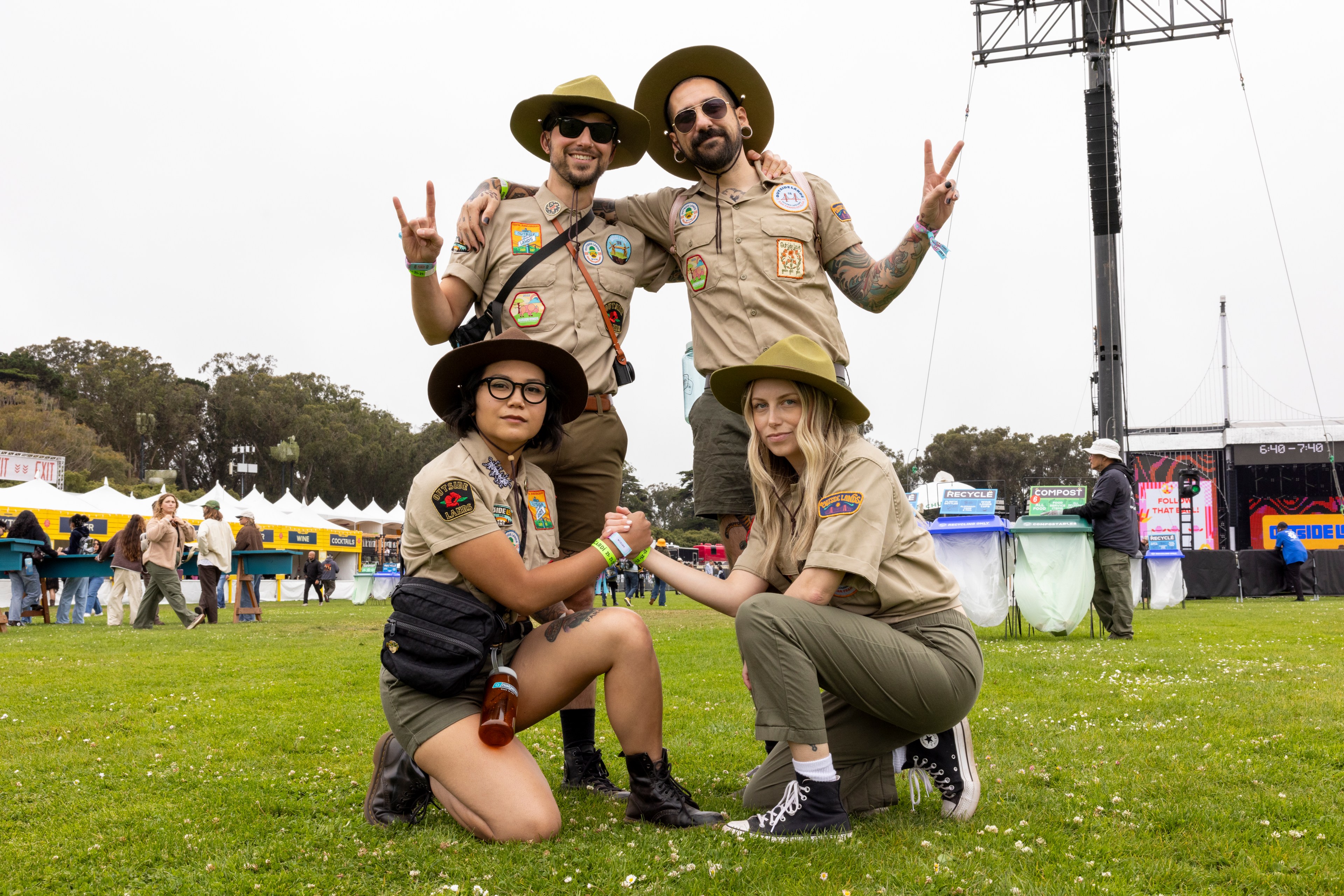 Four people in scout uniforms, two standing and two kneeling, pose on a grassy field at what appears to be an outdoor event with tents and a stage in the background.